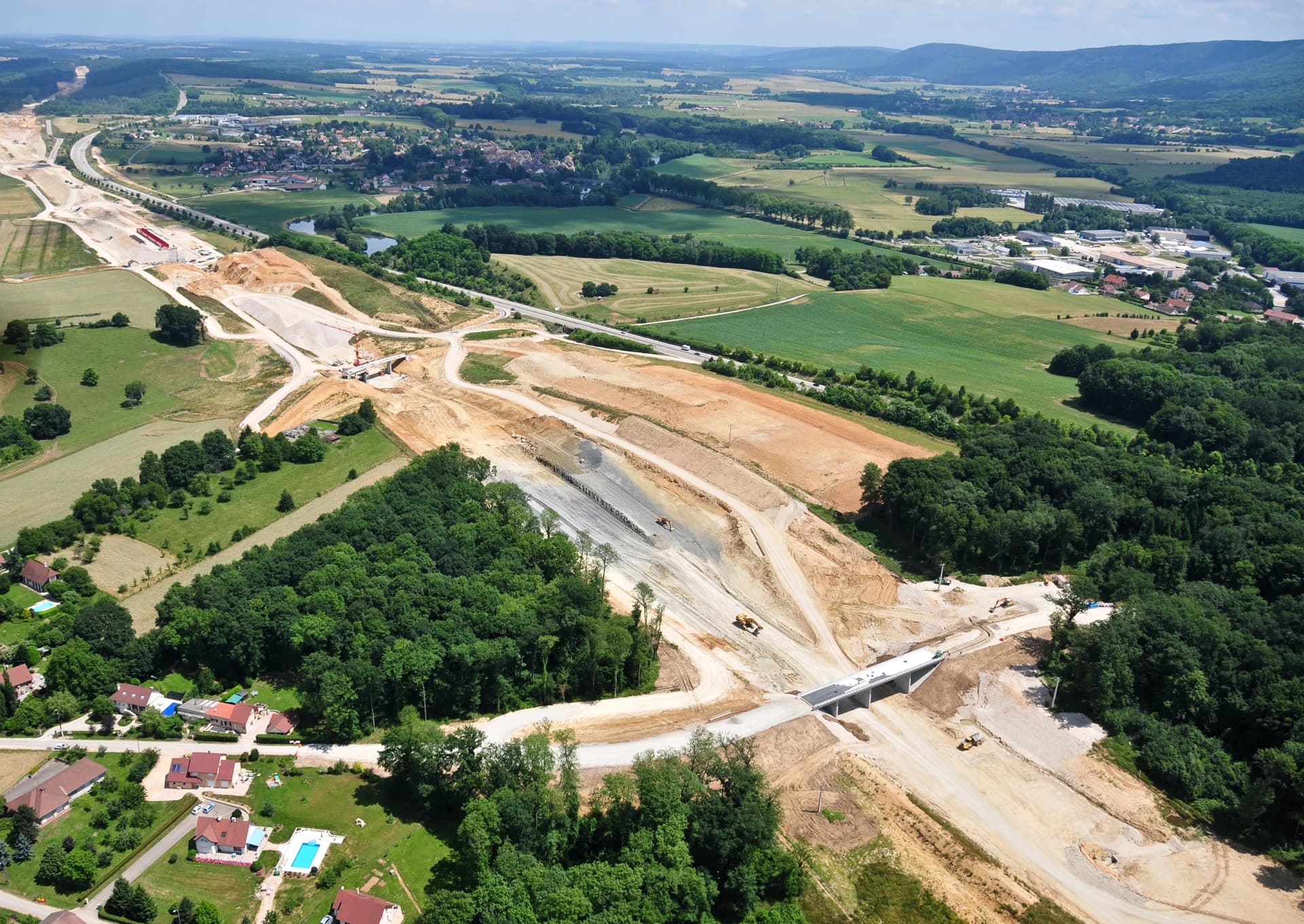 Vue aérienne d'une autoroute en construction, avec de larges tranchées de terre brune et des ponts traversant des champs verts, des forêts luxuriantes et des zones résidentielles, sous un ciel bleu.