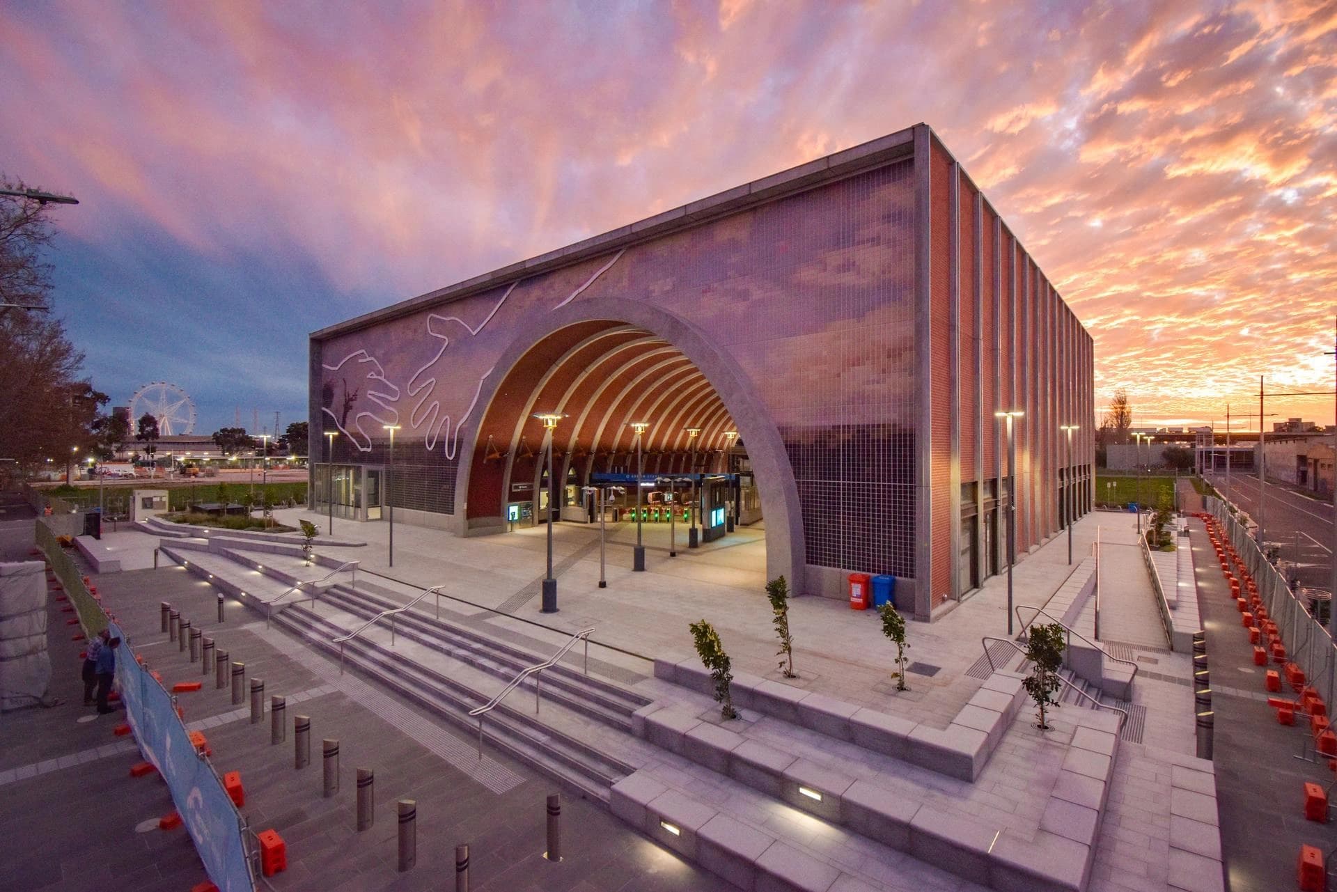 Façade d'une gare moderne avec une grande arche voûtée ornée d'une fresque de mains stylisées, sous un ciel de coucher de soleil aux teintes roses et oranges. Le parvis et les escaliers sont éclairés par des réverbères et des lumières intégrées, avec une grande roue visible au loin à gauche.
