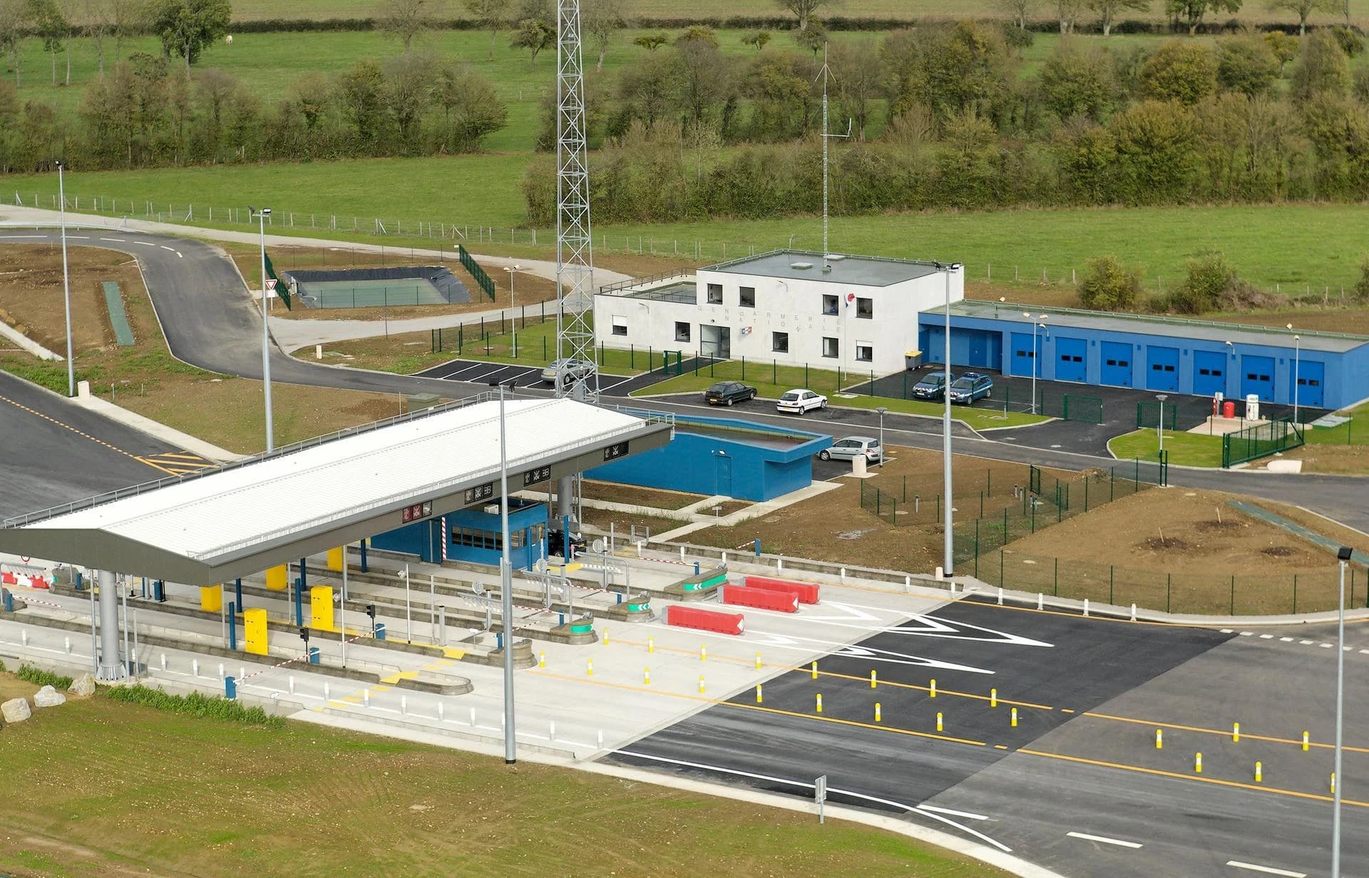 Une vue aérienne présente un complexe moderne de la Gendarmerie Nationale composé de bâtiments blancs et de garages bleus, jouxtant une station de péage ou de contrôle frontalier à plusieurs voies avec un grand auvent blanc et des routes balisées, le tout entouré de champs verdoyants.