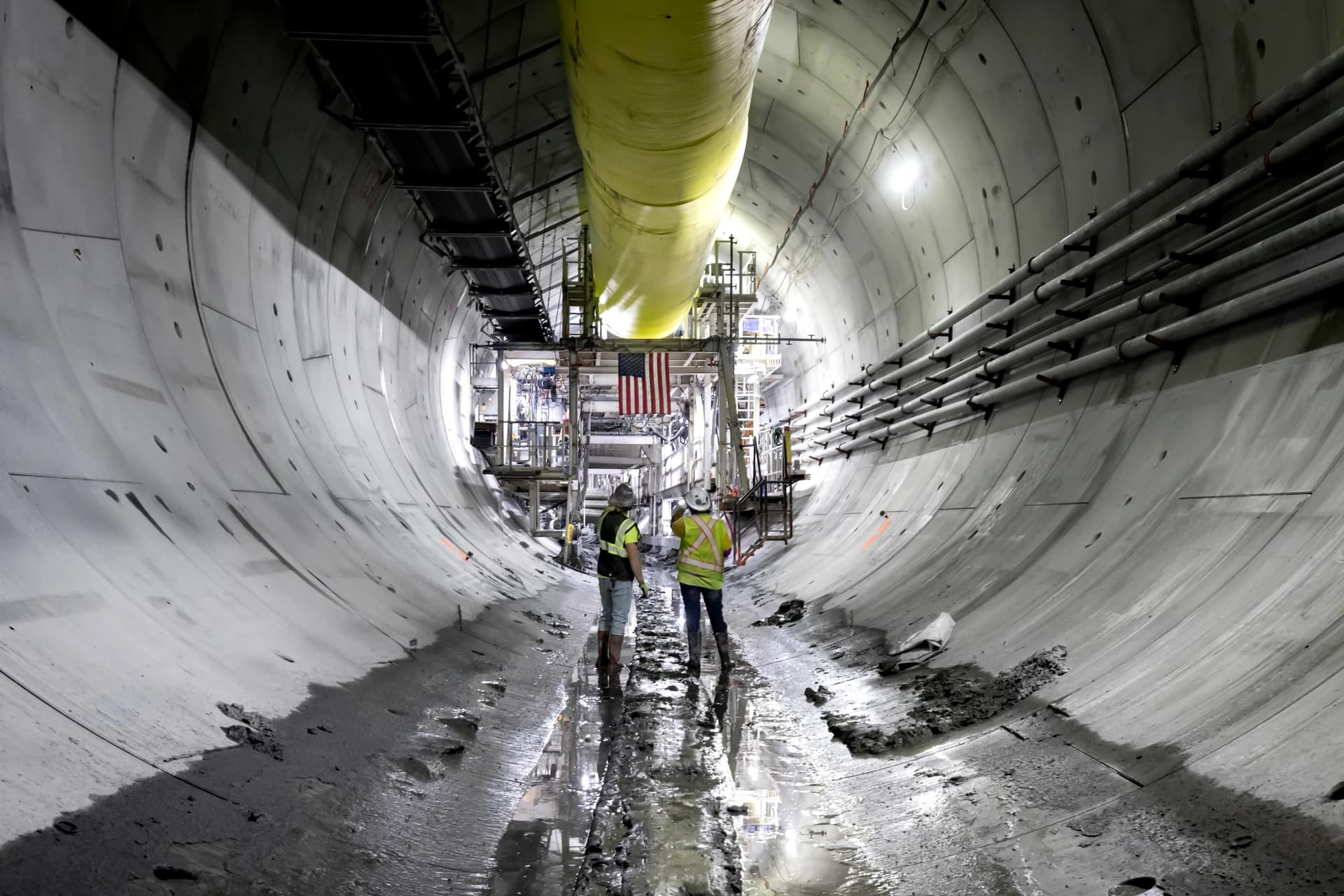 Deux ouvriers du bâtiment en gilets haute visibilité marchent dans un tunnel de béton gris en construction, avec un grand conduit jaune au plafond et un drapeau américain suspendu à l'équipement au fond. Le sol est humide et boueux.
