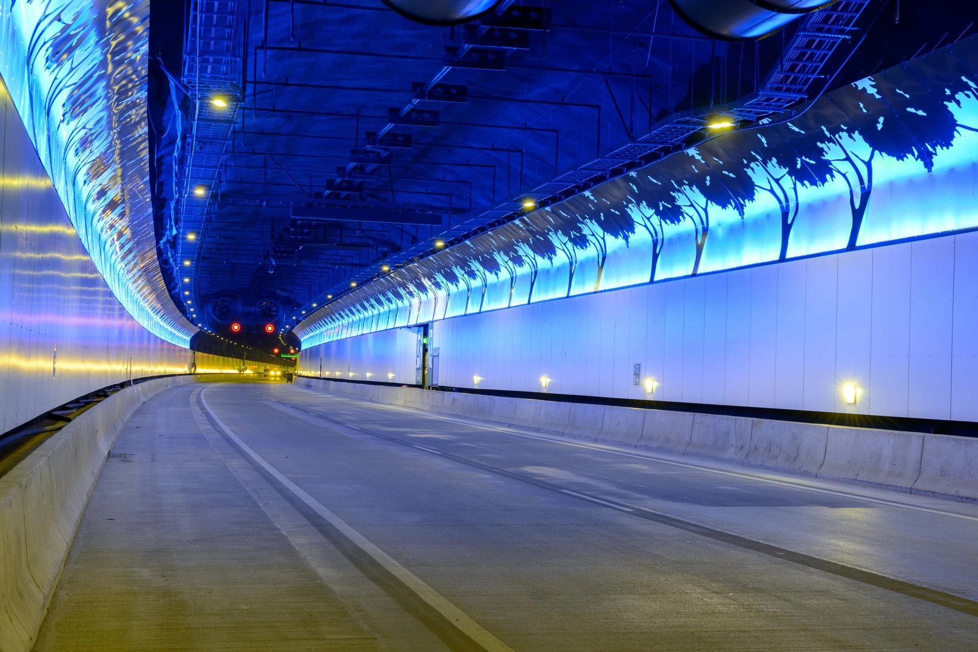 Un tunnel autoroutier moderne et vide est illuminé par un éblouissant affichage de lumière bleue avec des silhouettes d'arbres le long du mur de droite, contrastant avec l'éclairage jaune chaud du mur de gauche et de la route en béton incurvée.