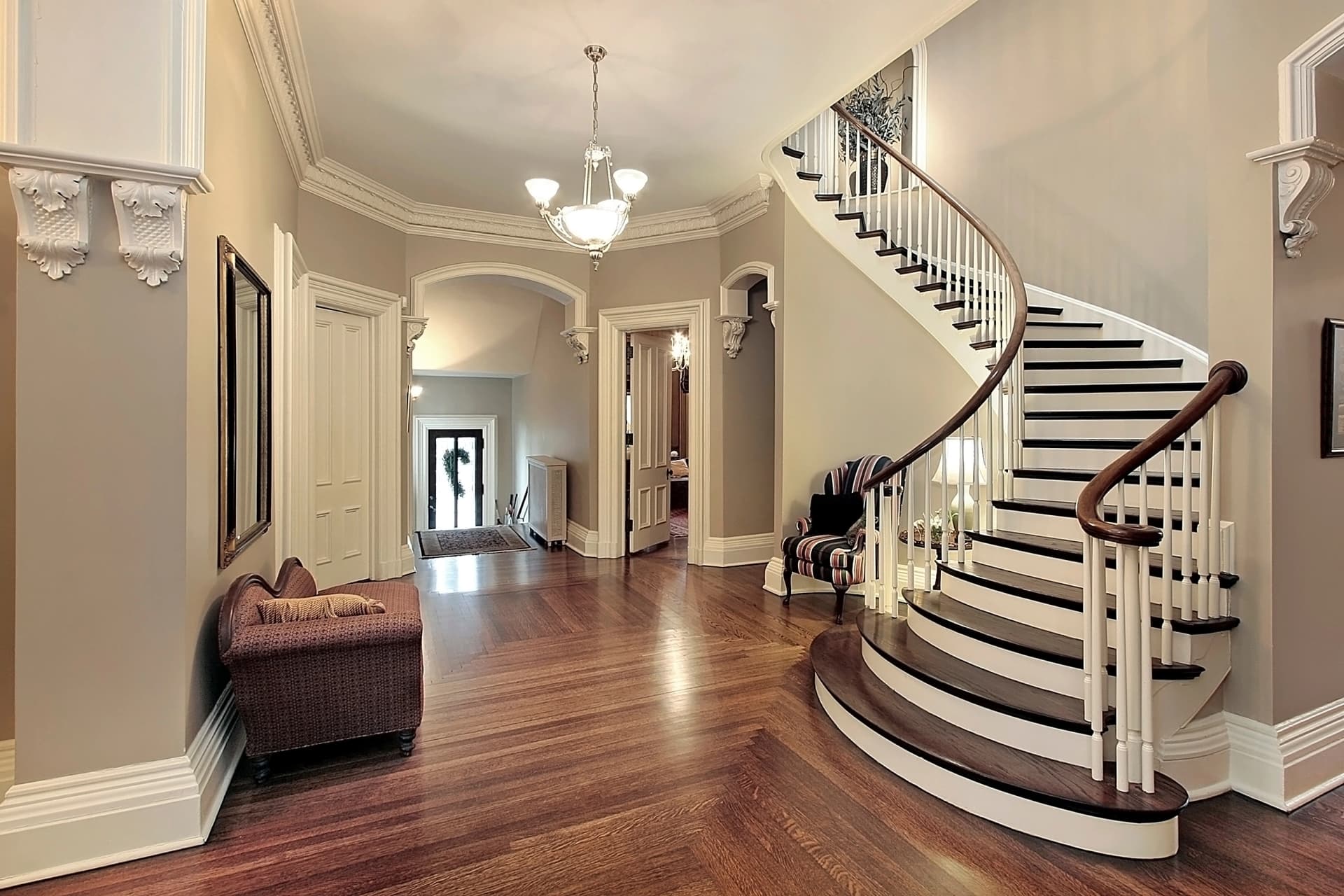 Un élégant foyer présente un grand escalier courbé avec des contremarches blanches et des marches en bois foncé, un sol en parquet à chevrons et des murs beiges clairs. Le décor comprend une garniture blanche ornée, un lustre central et des portes menant à d'autres pièces.