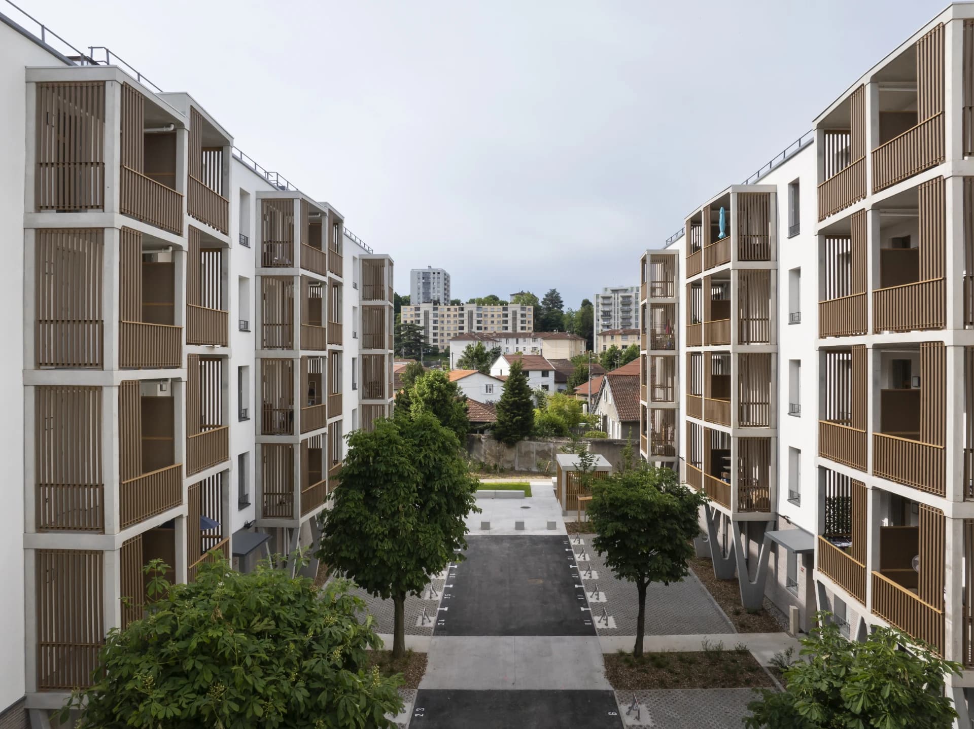Vue aérienne de deux immeubles résidentiels modernes et blancs à plusieurs étages, avec des balcons à écrans de lattes de bois clair, encadrant une allée pavée bordée d'arbres. En arrière-plan, un quartier urbain mixte se déploie sous un ciel nuageux et lumineux.