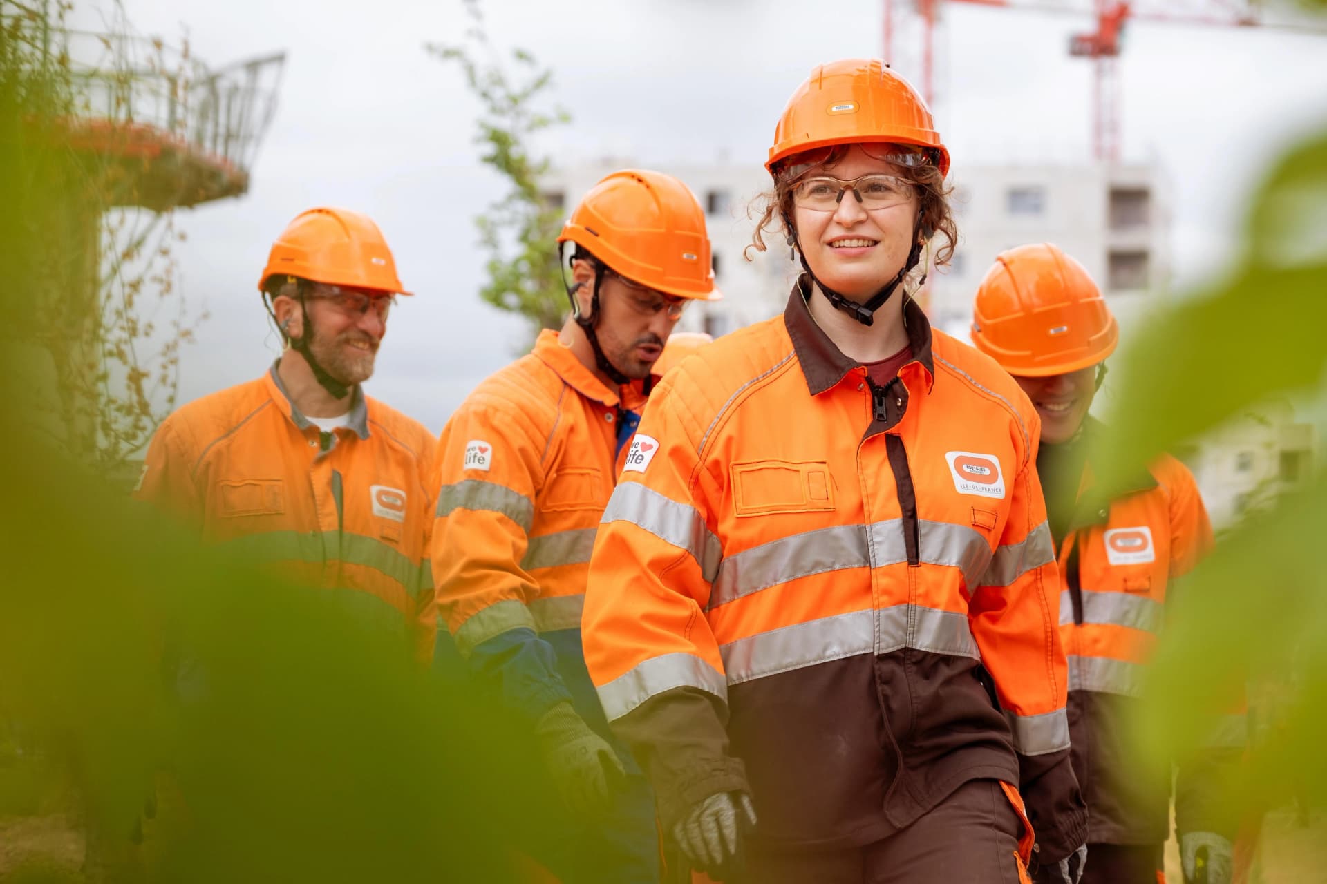 Une femme souriante en casque orange, lunettes de sécurité et veste de travail orange et marron haute visibilité est au premier plan, encadrée par un feuillage vert flou. Derrière elle, trois autres ouvriers en tenue de sécurité orange similaire et une grue de chantier rouge sont visibles sur un site de construction.