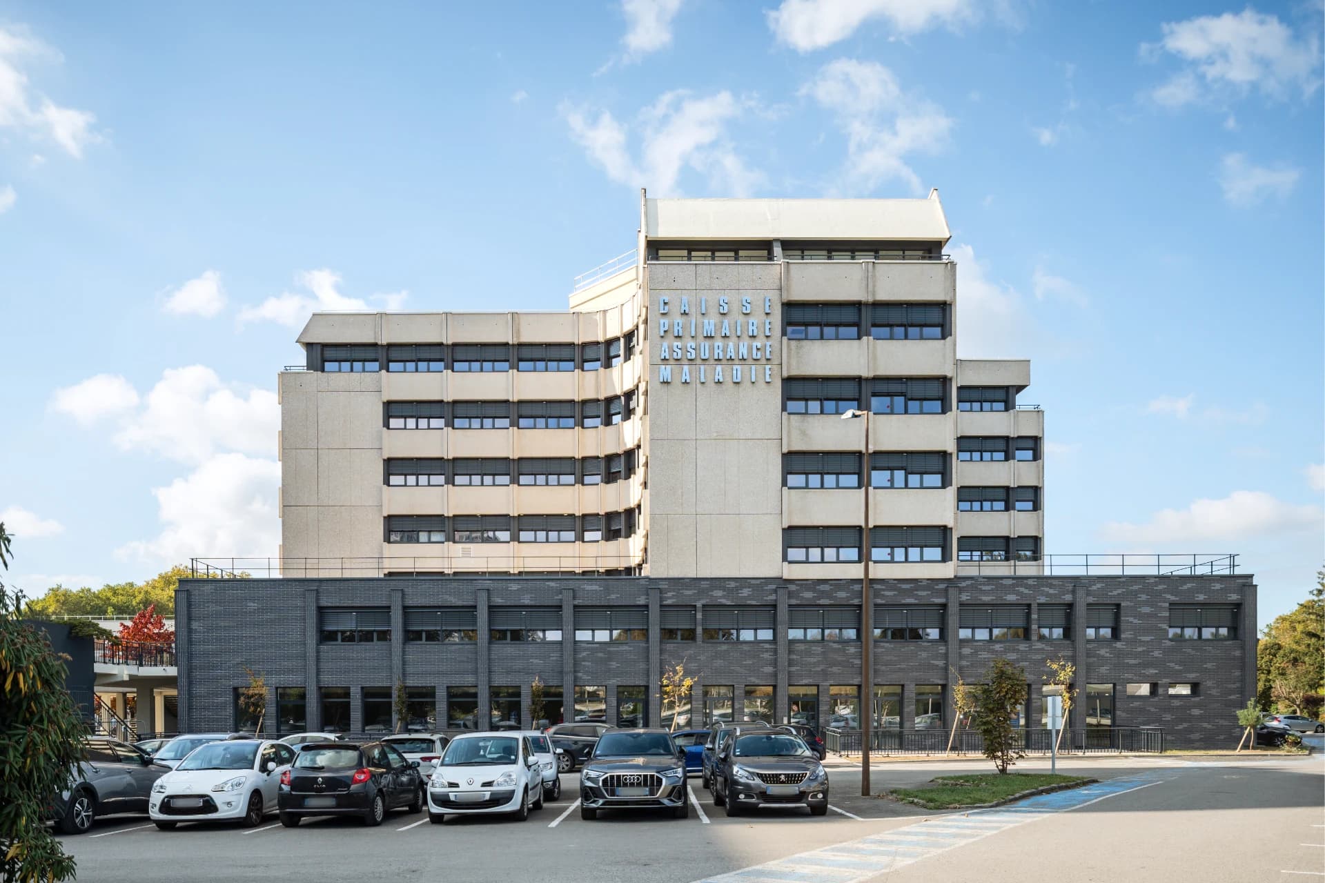Un grand immeuble de la Caisse Primaire Assurance Maladie, avec des lettres bleues verticales sur sa façade supérieure gris clair et une base en briques sombres, est visible sous un ciel bleu avec un grand parking rempli de voitures au premier plan.
