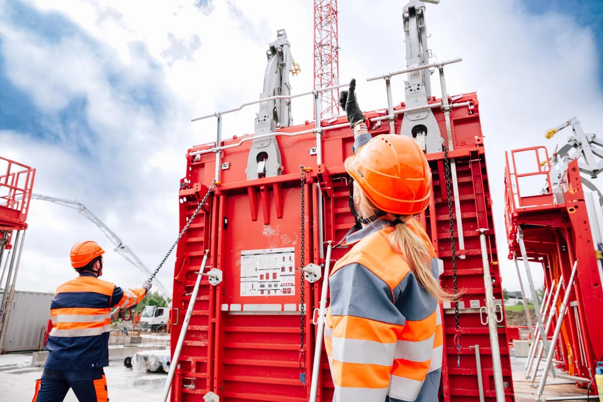 Deux ouvriers du bâtiment en casque et veste orange supervisent d'imposantes structures de coffrage rouges sur un chantier, l'un pointant vers le haut sous un ciel nuageux.