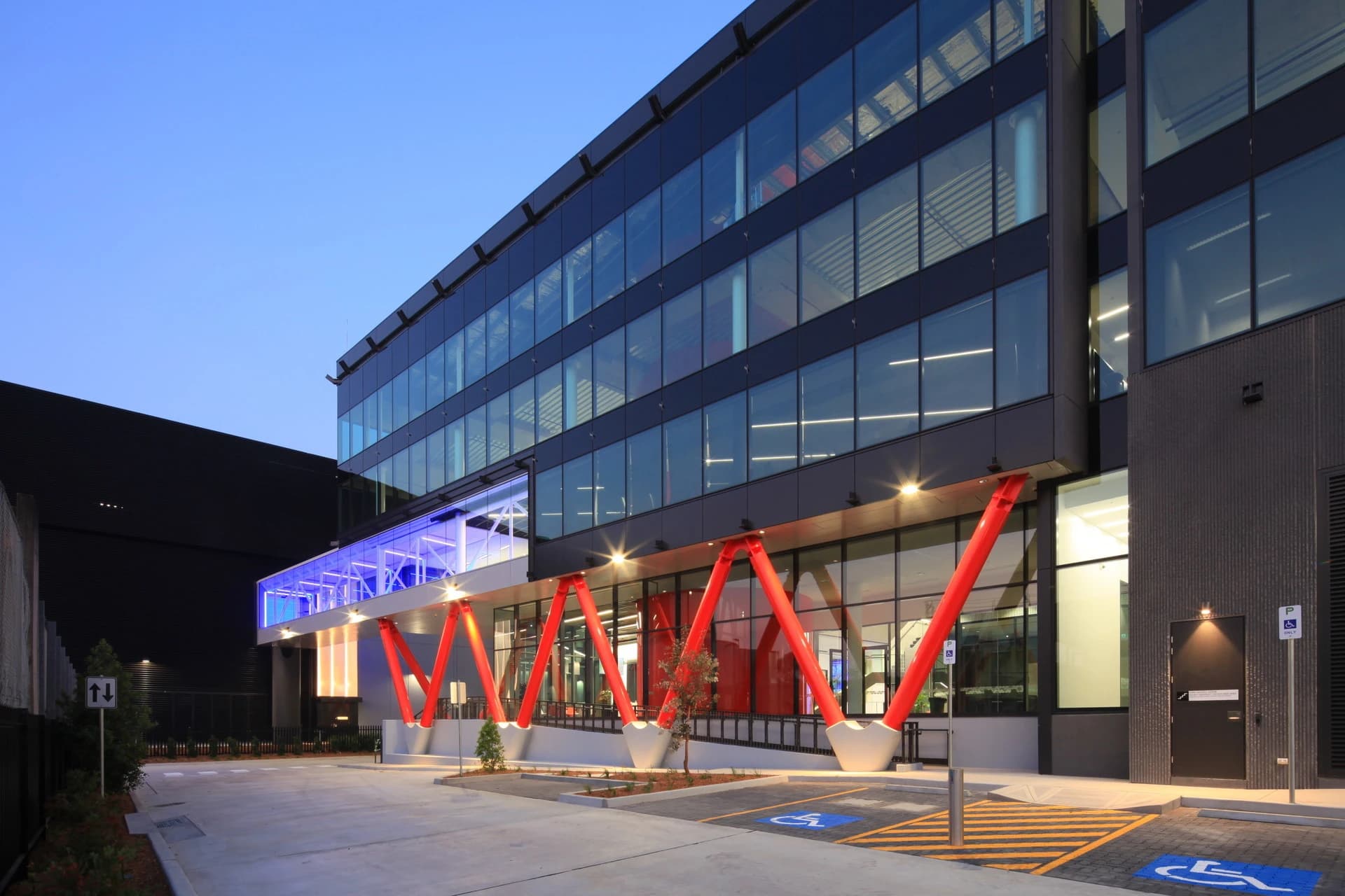Un bâtiment moderne à plusieurs étages avec une façade en verre sombre, soutenu par de grandes poutres diagonales rouges au rez-de-chaussée. Une passerelle d'entrée est éclairée en bleu, avec des places de parking accessibles en premier plan.