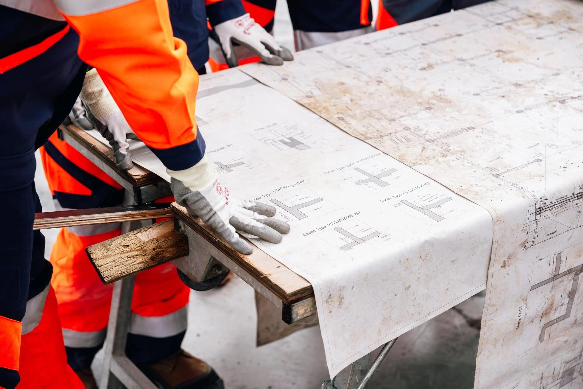 Des ouvriers en gants et tenues de sécurité orange et bleu marine examinent un grand plan de construction détaillé, légèrement sali, posé sur une surface en bois.