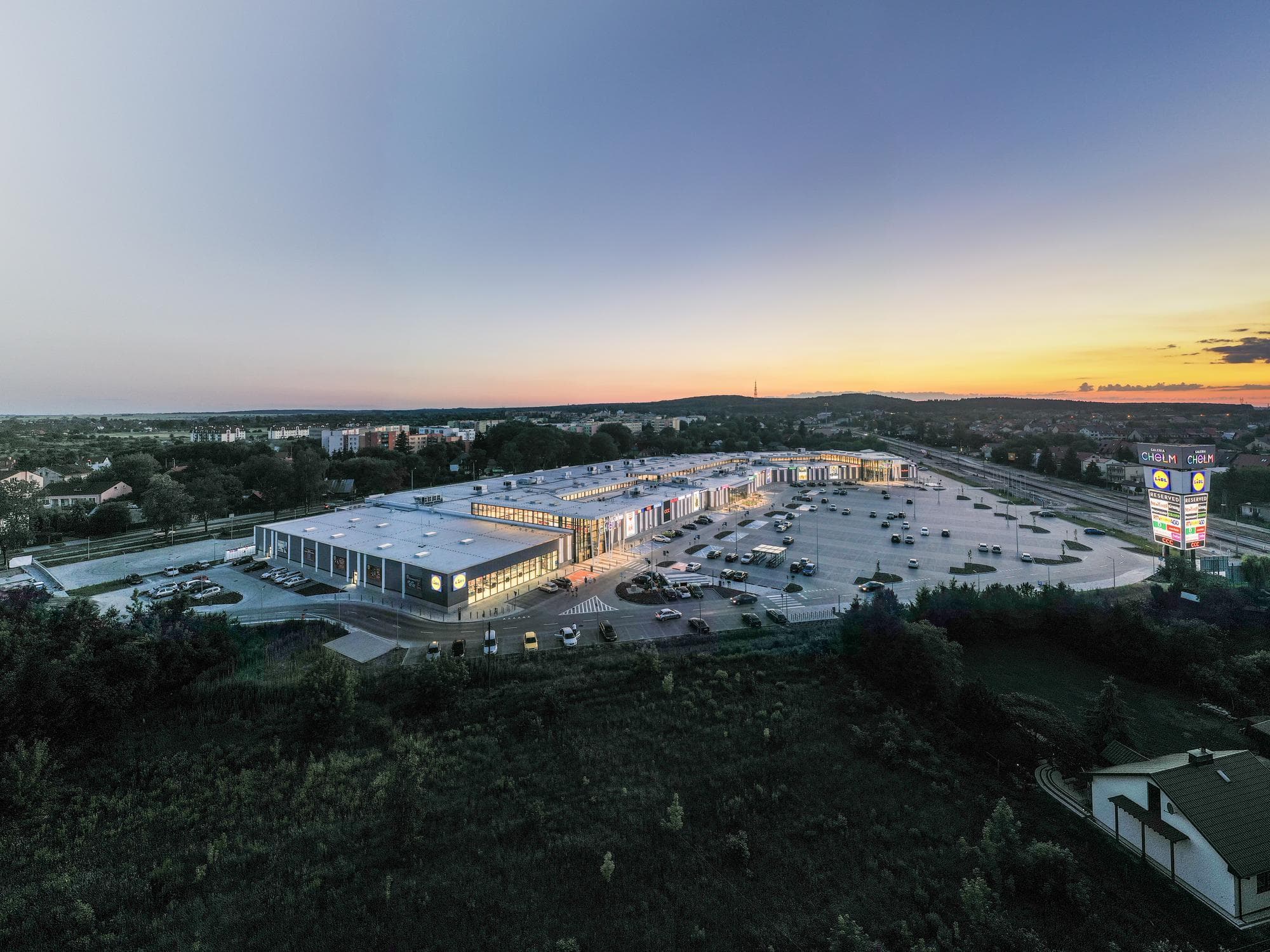 Vue aérienne d'un grand centre commercial moderne et de son vaste parking, brillamment éclairés au crépuscule. Le ciel passe du bleu à l'orange au-dessus d'un paysage de verdure luxuriante et d'une ville lointaine, avec des enseignes lumineuses comme Lidl et Galeria Chełm.