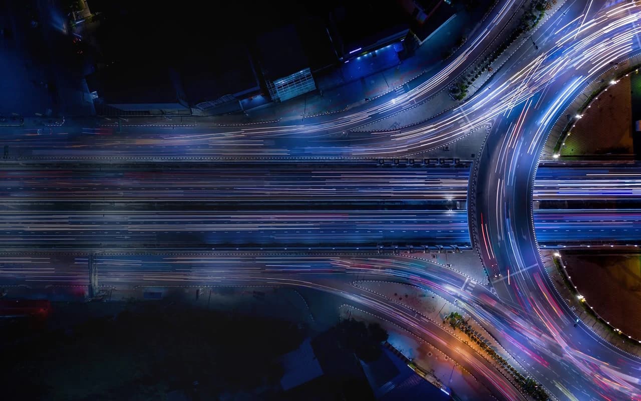 Vue aérienne de nuit d'un échangeur routier avec des traînées lumineuses de véhicules, symbolisant le mouvement et la connectivité urbaine.