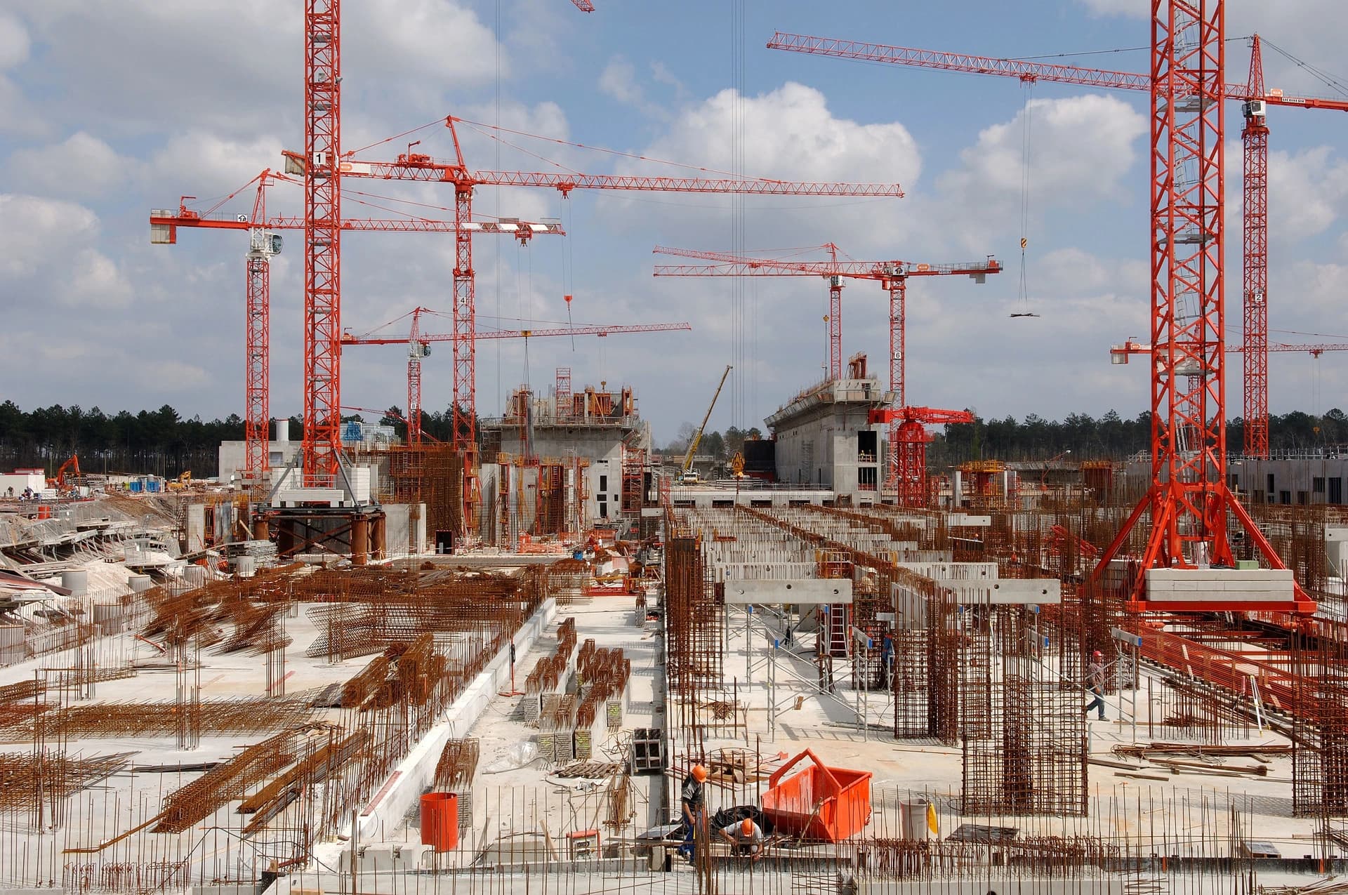 Un vaste chantier de construction présente de nombreuses grues rouges s'élevant au-dessus de structures en béton armé et d'abondantes armatures métalliques rouillées, sous un ciel nuageux et une ligne d'arbres en arrière-plan.