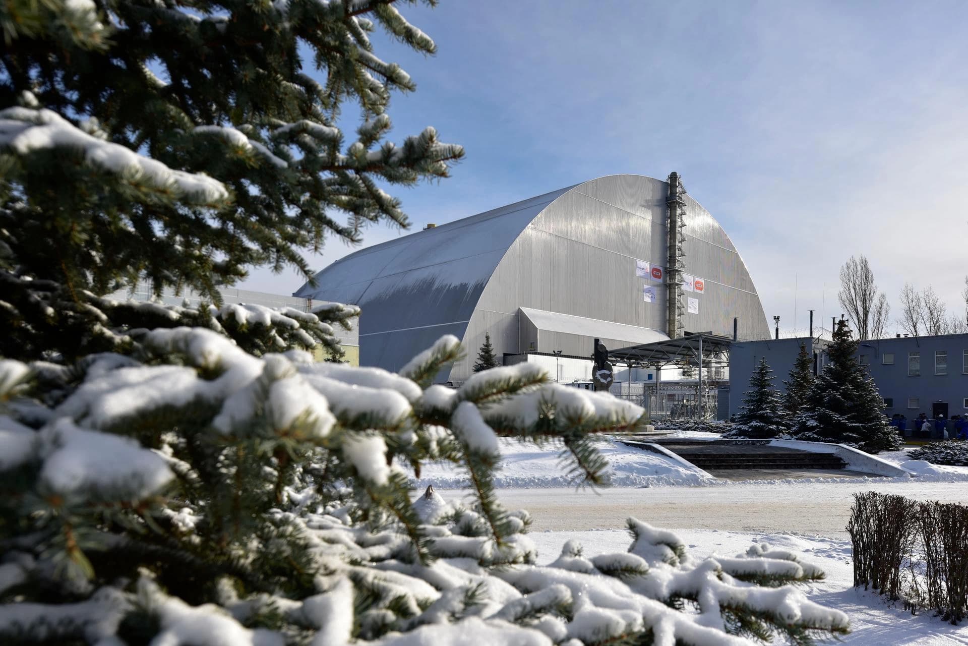 La massive arche de confinement grise de Tchernobyl est visible sous un ciel bleu clair, encadrée par des branches de sapin chargées de neige au premier plan, dans un paysage hivernal.