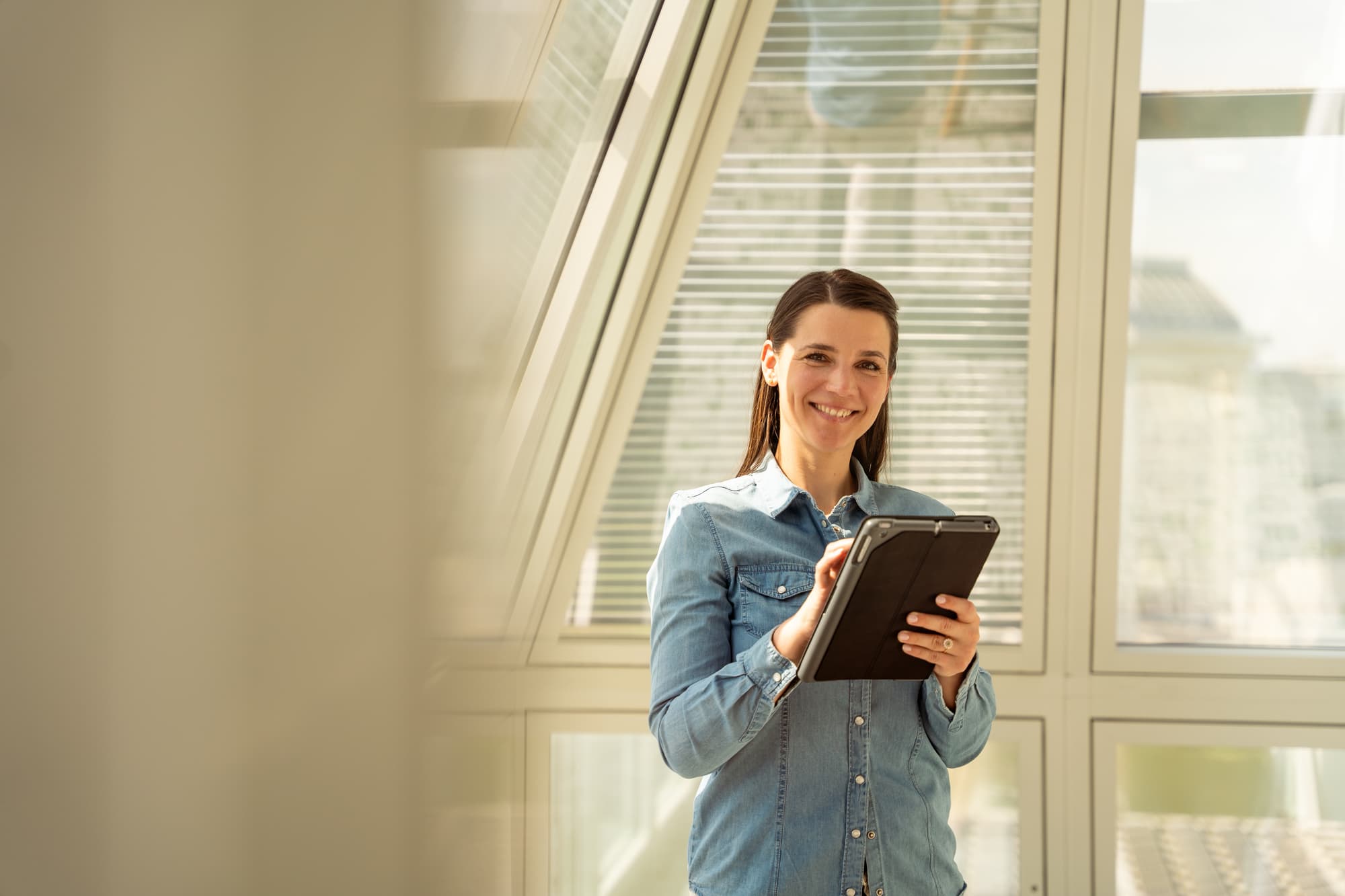 Une femme souriante, vêtue d'une chemise en jean bleue, tient une tablette devant de grandes fenêtres lumineuses avec des stores, offrant une vue sur la ville.