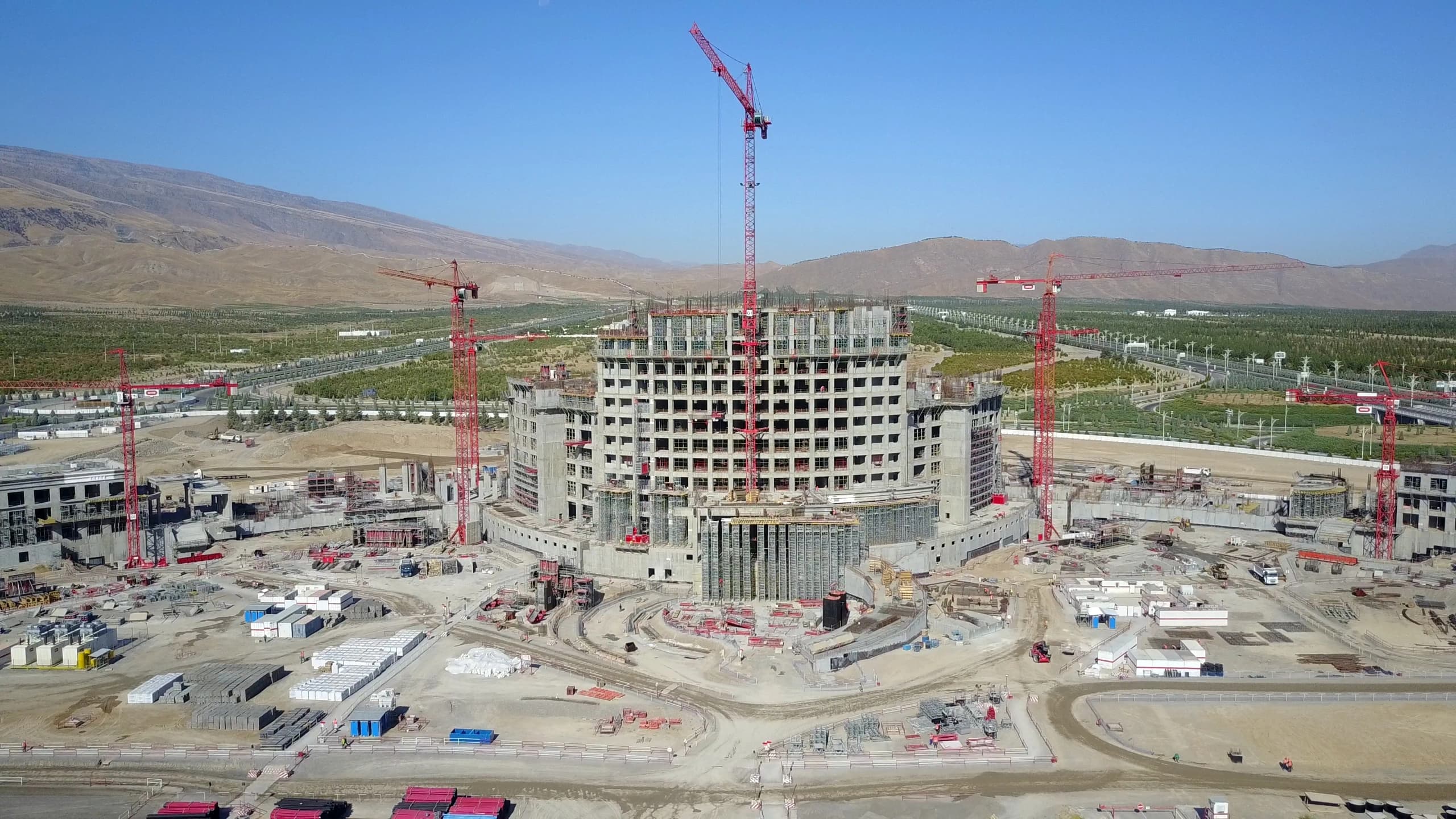 Vue aérienne d'un grand bâtiment gris en béton en construction, cerné par de multiples grues rouges et un vaste chantier. L'ensemble est situé dans un paysage aride avec des montagnes ocres en arrière-plan et des zones verdoyantes irriguées sous un ciel bleu clair.