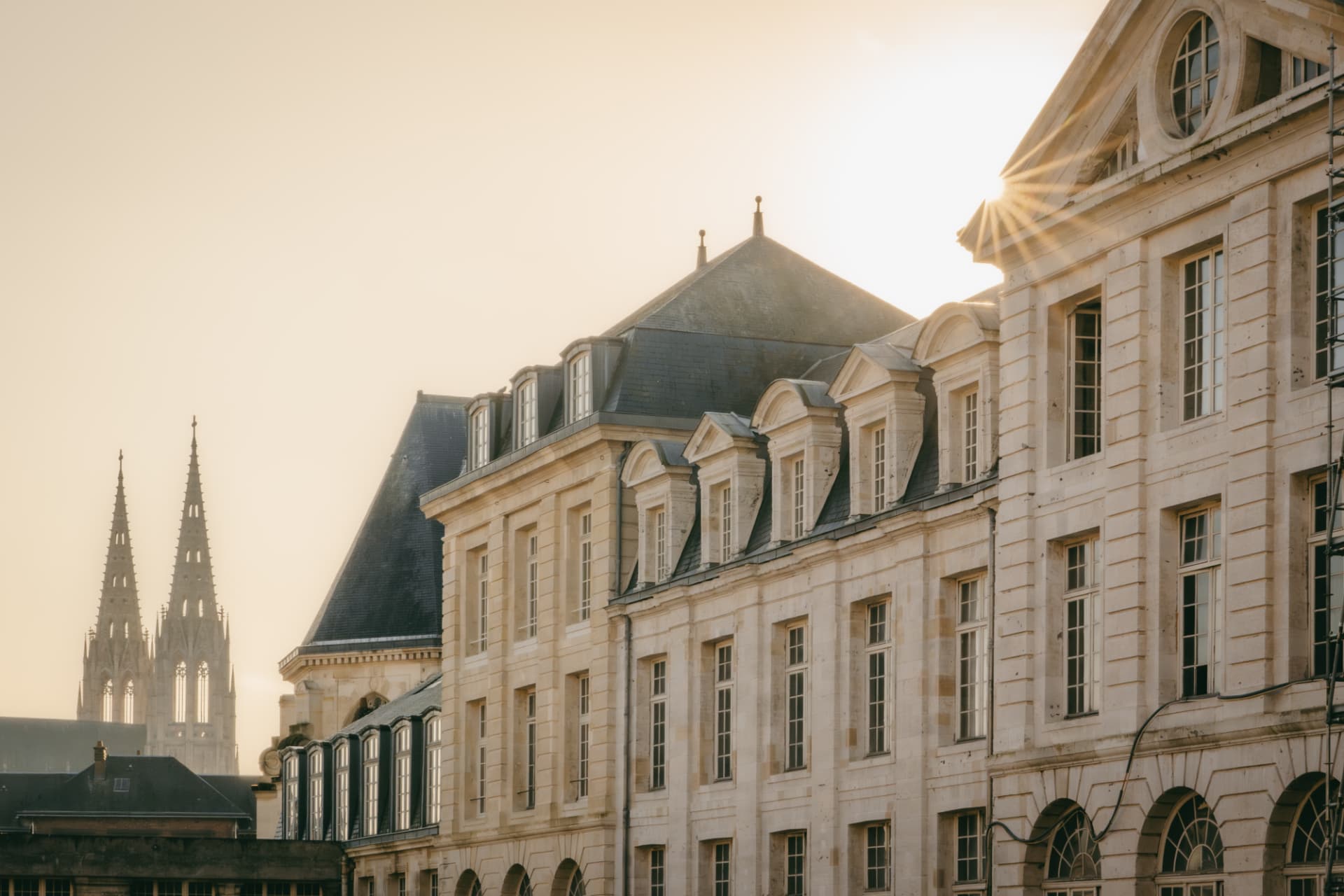 Façades classiques en pierre beige avec des toits en ardoise sombre, illuminées par un soleil rasant créant des rayons dorés, avec deux clochers de cathédrale gothique en arrière-plan sous un ciel clair.