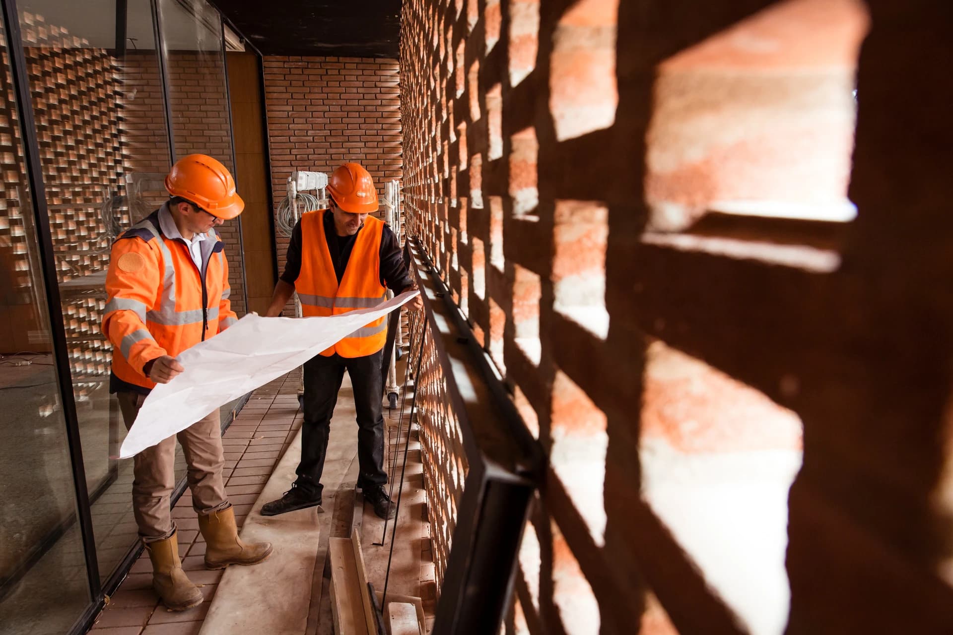 Deux ouvriers du bâtiment en tenue de sécurité orange et casques examinent un grand plan architectural à l'intérieur d'un bâtiment en construction, où la lumière du soleil crée des motifs d'ombre saisissants sur un mur de briques.