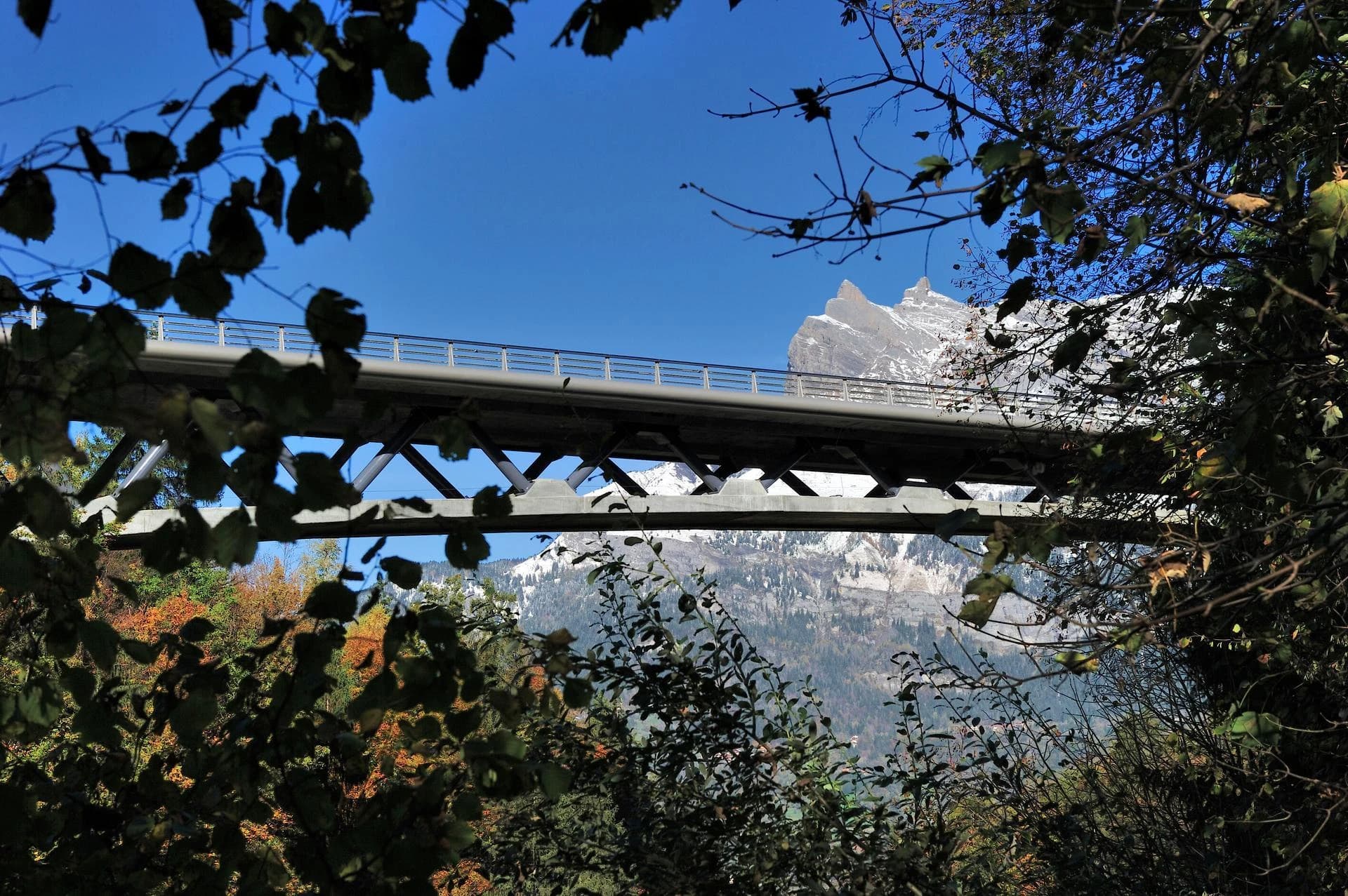 Un robuste pont gris s'étend sur une vallée, encadré par un feuillage d'automne aux couleurs vives, avec d'imposantes montagnes enneigées sous un ciel bleu clair en arrière-plan.