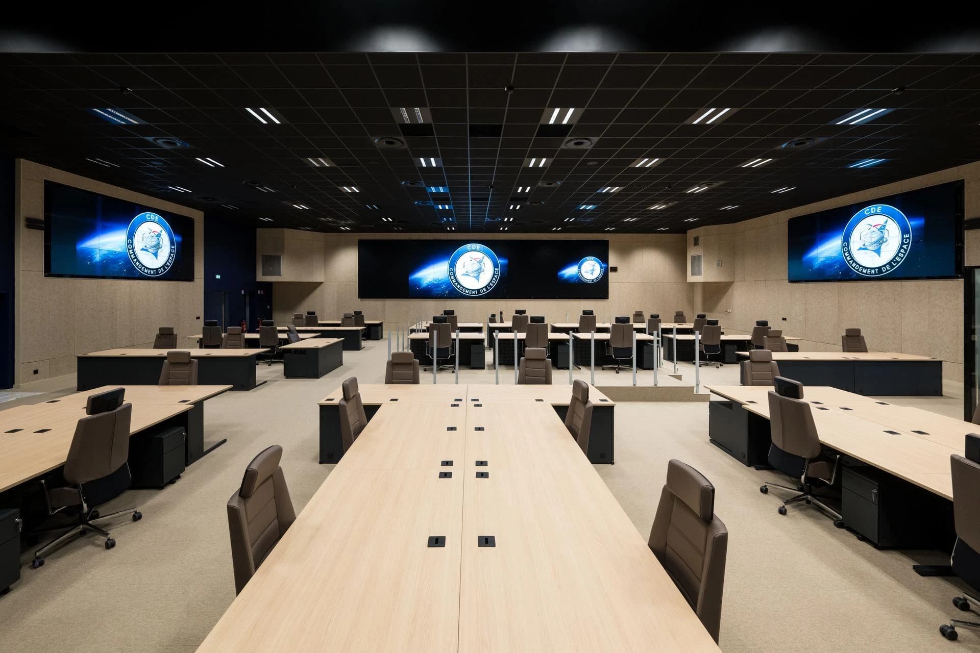 Vaste salle de commande moderne avec des rangées de bureaux en bois clair et de chaises marron, faisant face à de grands écrans affichant le logo "CDE COMMANDEMENT DE L'ESPACE" sur un fond spatial bleu. L'espace est caractérisé par des murs beiges et un plafond noir quadrillé de lumières.