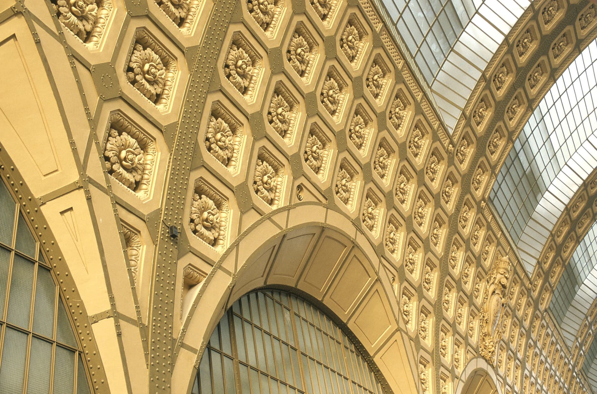 Plafond et murs supérieurs d'un grand édifice présentant un motif répétitif de panneaux hexagonaux dorés, chacun orné d'un motif floral sculpté, intégrés dans une structure métallique rivetée. De grandes baies vitrées et des verrières voûtées diffusent la lumière naturelle, créant une ambiance majestueuse.