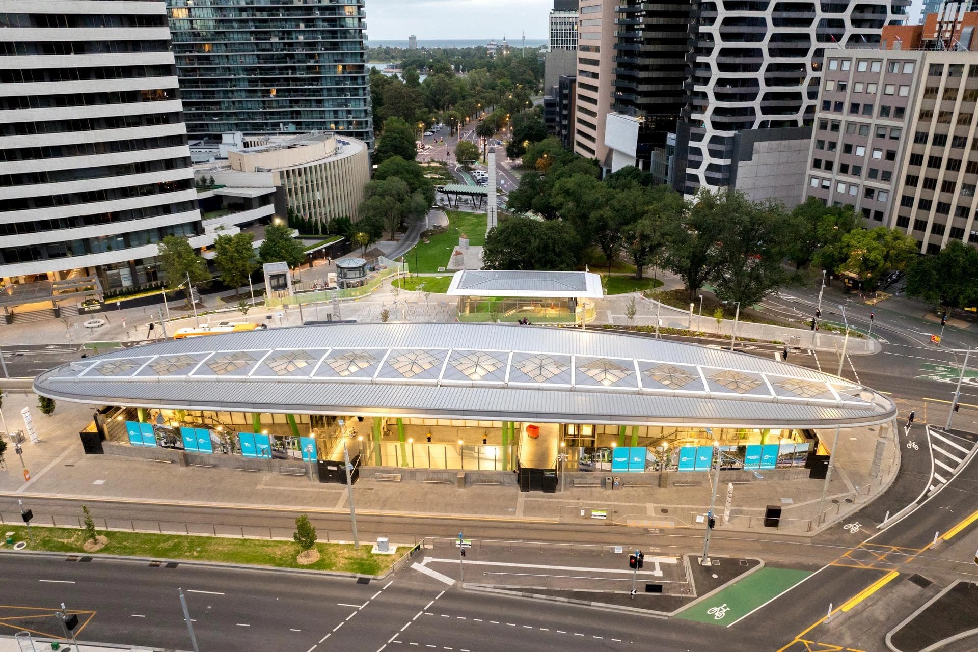 Une vue aérienne révèle une station de transport public moderne et allongée, avec un toit gris incurvé et des intérieurs lumineux orange-jaune visibles à travers des panneaux vitrés décorés de publicités bleues. Elle est située au cœur d'un environnement urbain dense, entourée de gratte-ciel, de routes avec pistes cyclables vertes, et d'espaces verts.