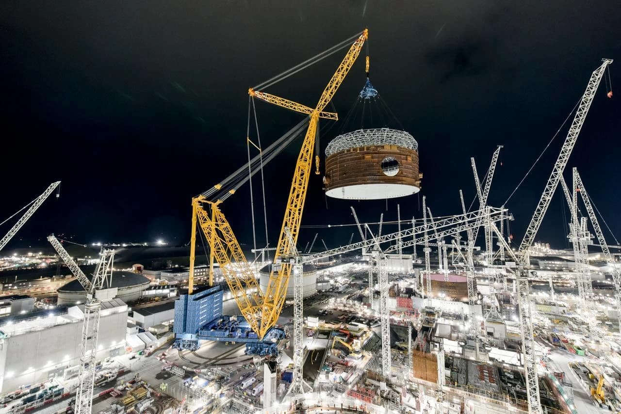 Grande grue chenille jaune levant une section cylindrique marron et blanche au-dessus d'un vaste chantier de construction brillamment éclairé la nuit, entouré de nombreuses autres grues et de structures industrielles en cours d'assemblage.