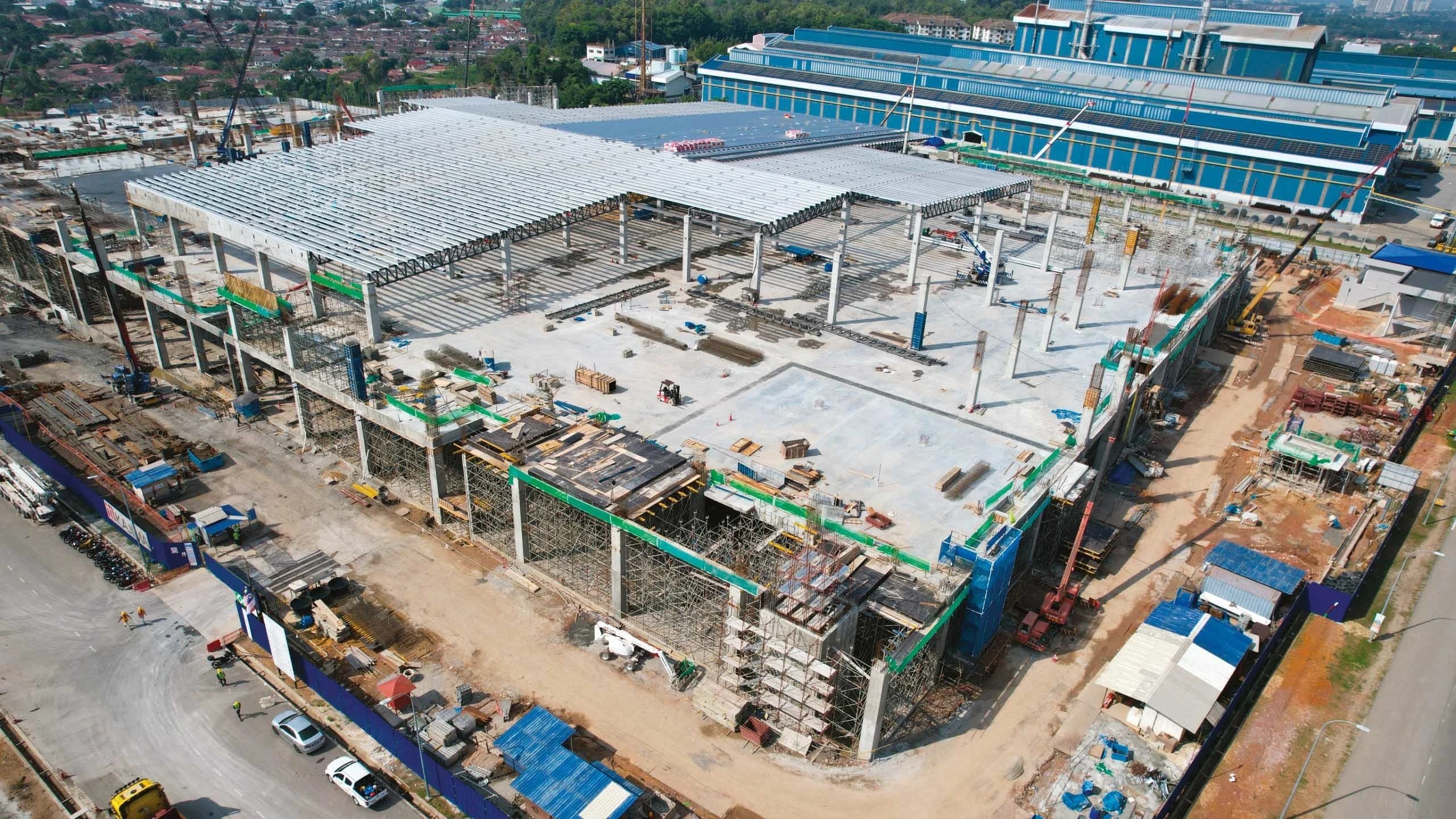 Vue aérienne d'un vaste chantier de construction industrielle montrant une grande structure en béton et acier en cours de montage avec des toits partiels. Des équipements et matériaux sont dispersés, avec de grands bâtiments industriels bleus et un paysage urbain en arrière-plan.