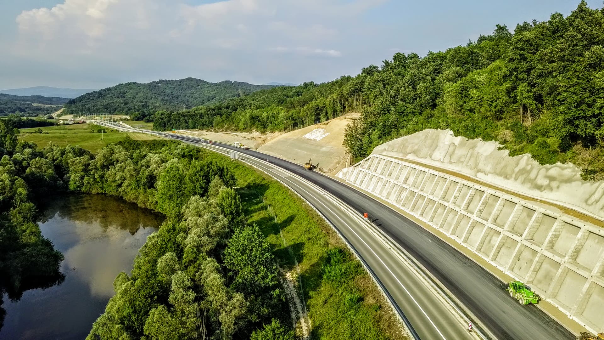 Vue aérienne d'une autoroute en construction avec de l'asphalte frais et des murs de soutènement en béton, traversant un paysage verdoyant composé de forêts denses, de champs et d'une rivière. Plusieurs engins de chantier, dont un vert vif, sont visibles sur la route et les talus, sous un ciel partiellement nuageux.