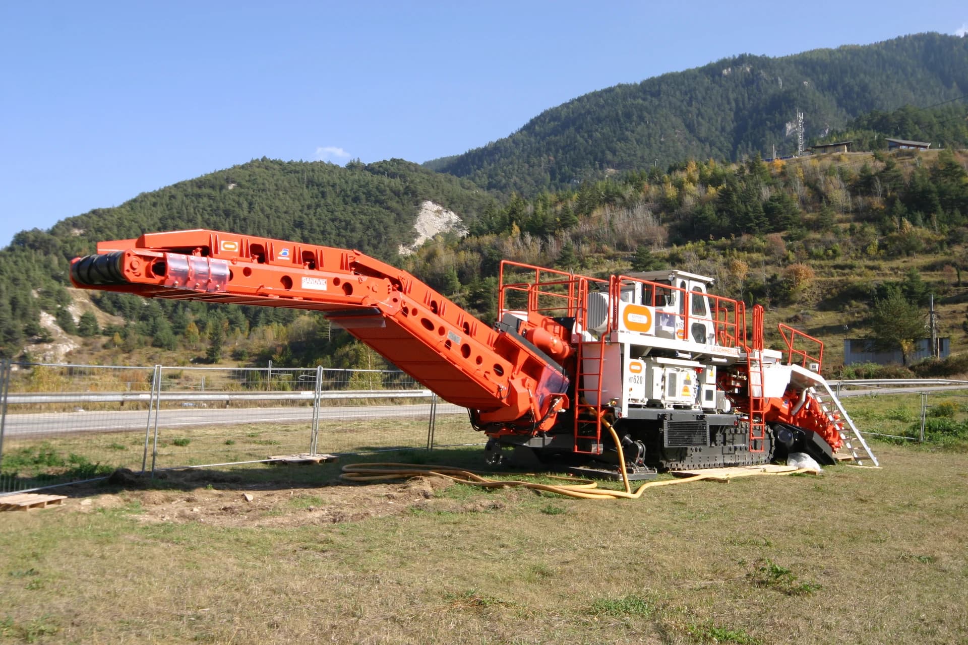 Une concasseuse mobile Sandvik MT620 orange et blanche sur chenilles, avec un long bras convoyeur, est garée sur de l'herbe sèche devant une route et une montagne boisée sous un ciel bleu clair.
