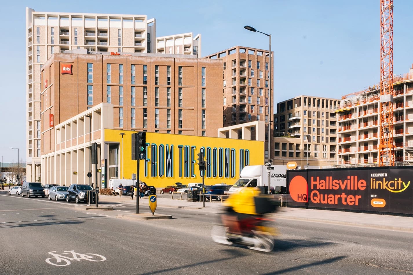A vibrant urban street scene with modern brick and concrete buildings, a bright yellow wall displaying "FROM THE GROUND UP," and a blurred delivery rider on a motorcycle passing a construction site with an orange crane and development signage.