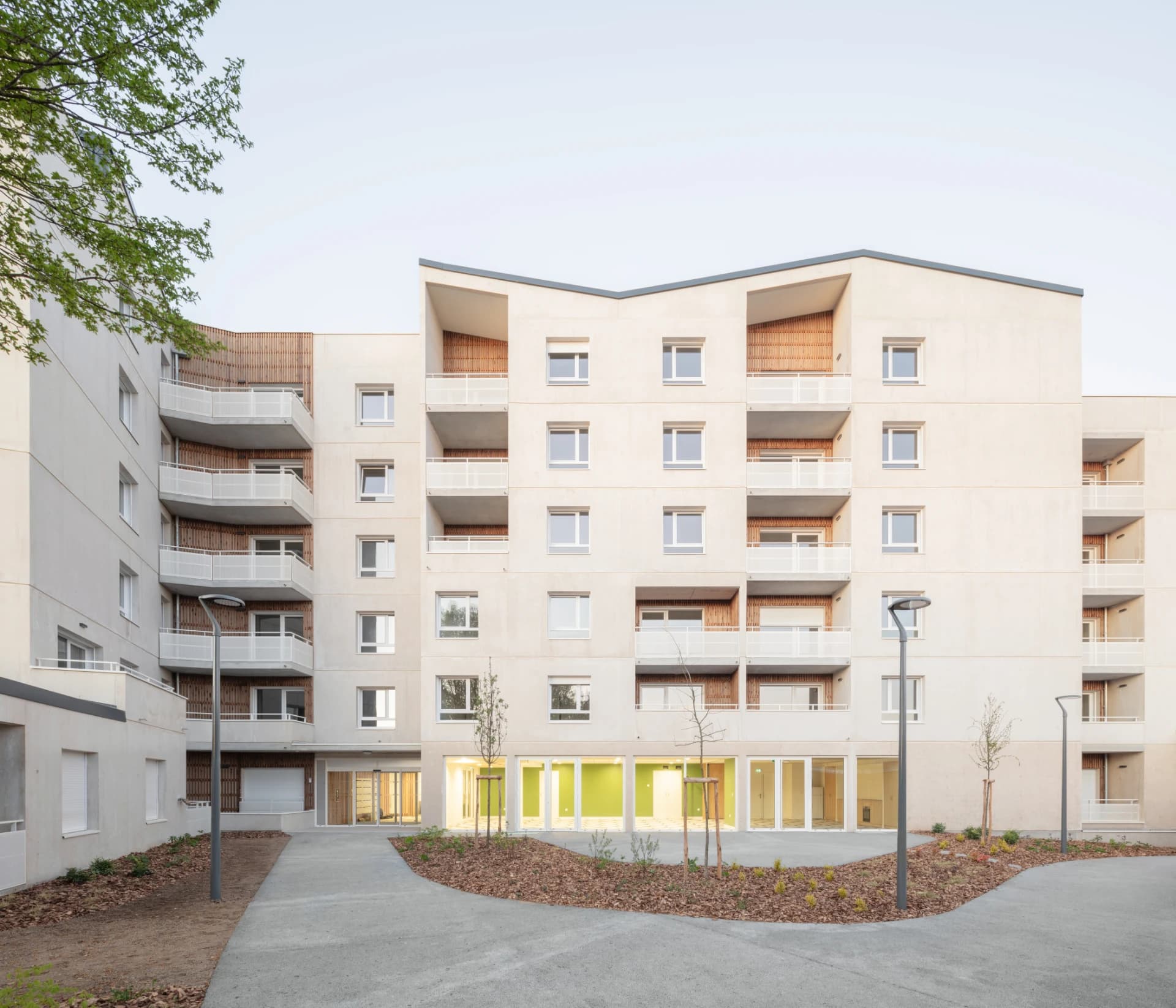 Grand immeuble résidentiel moderne beige clair avec balcons blancs et accents en bois, s'ouvrant sur une cour aménagée de chemins sinueux, d'arbustes et de fenêtres de rez-de-chaussée éclairées en vert.