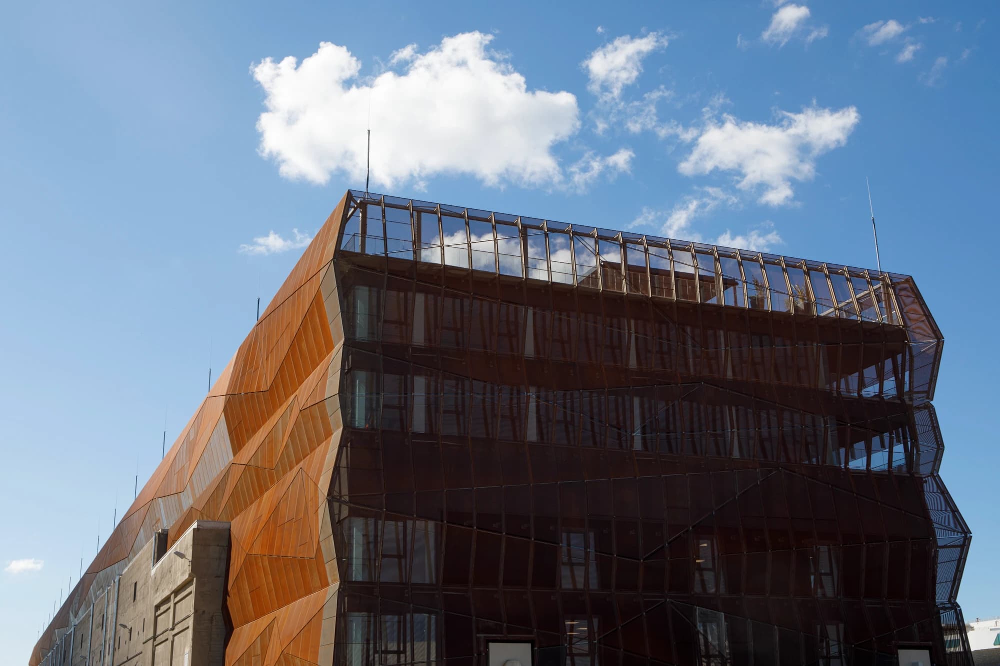 Bâtiment moderne à la façade complexe de panneaux géométriques orange rouille et brun foncé, avec un dernier étage vitré, sous un ciel bleu clair parsemé de nuages blancs.