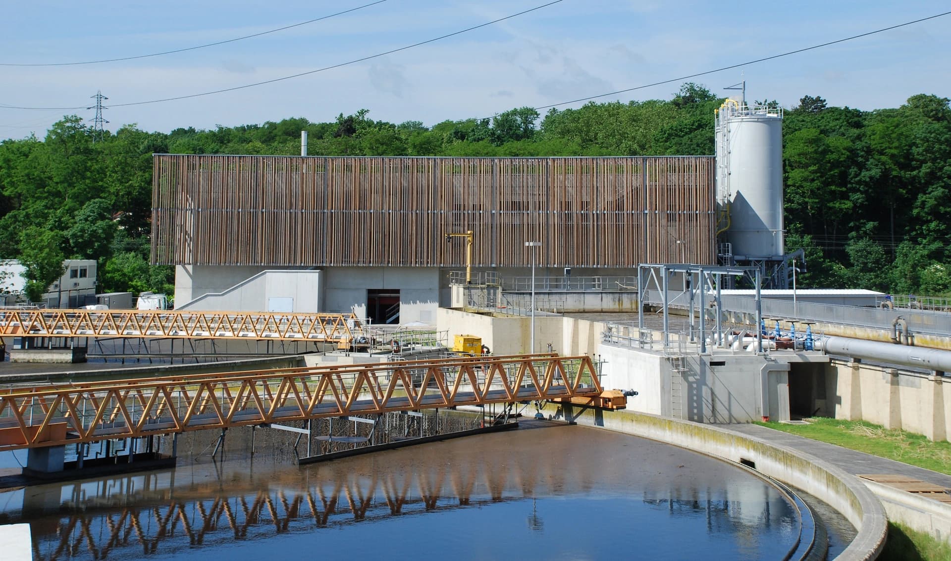 Une station d'épuration moderne avec un bâtiment principal à façade de lattes de bois verticales, un grand bassin de décantation circulaire reflétant des passerelles en treillis orange, et un silo gris, sous un ciel bleu entouré d'arbres verts.