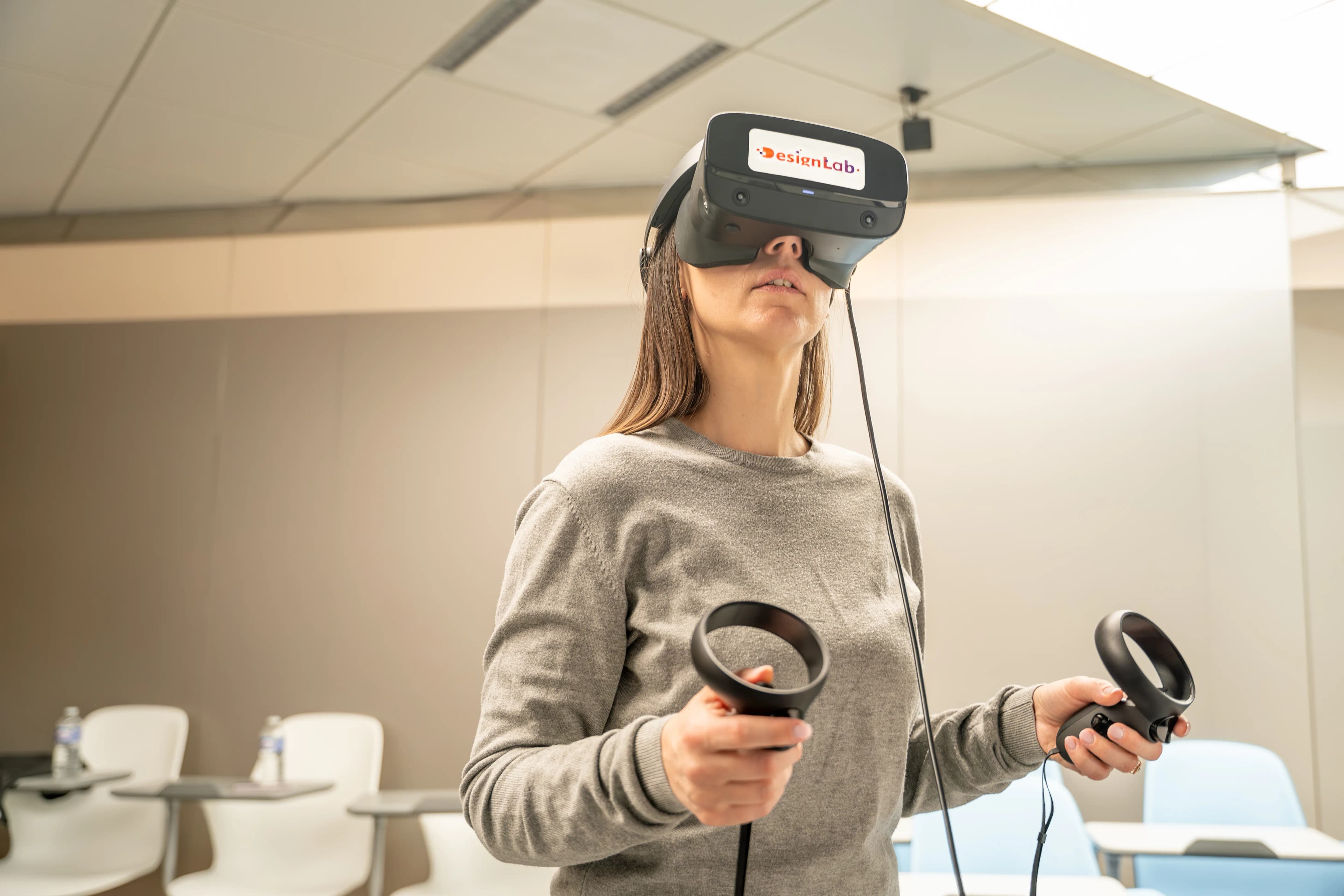 Une femme portant un casque de réalité virtuelle noir "DesignLab" et tenant deux contrôleurs est engagée dans une expérience virtuelle, dans une pièce moderne et lumineuse avec des chaises claires en arrière-plan.