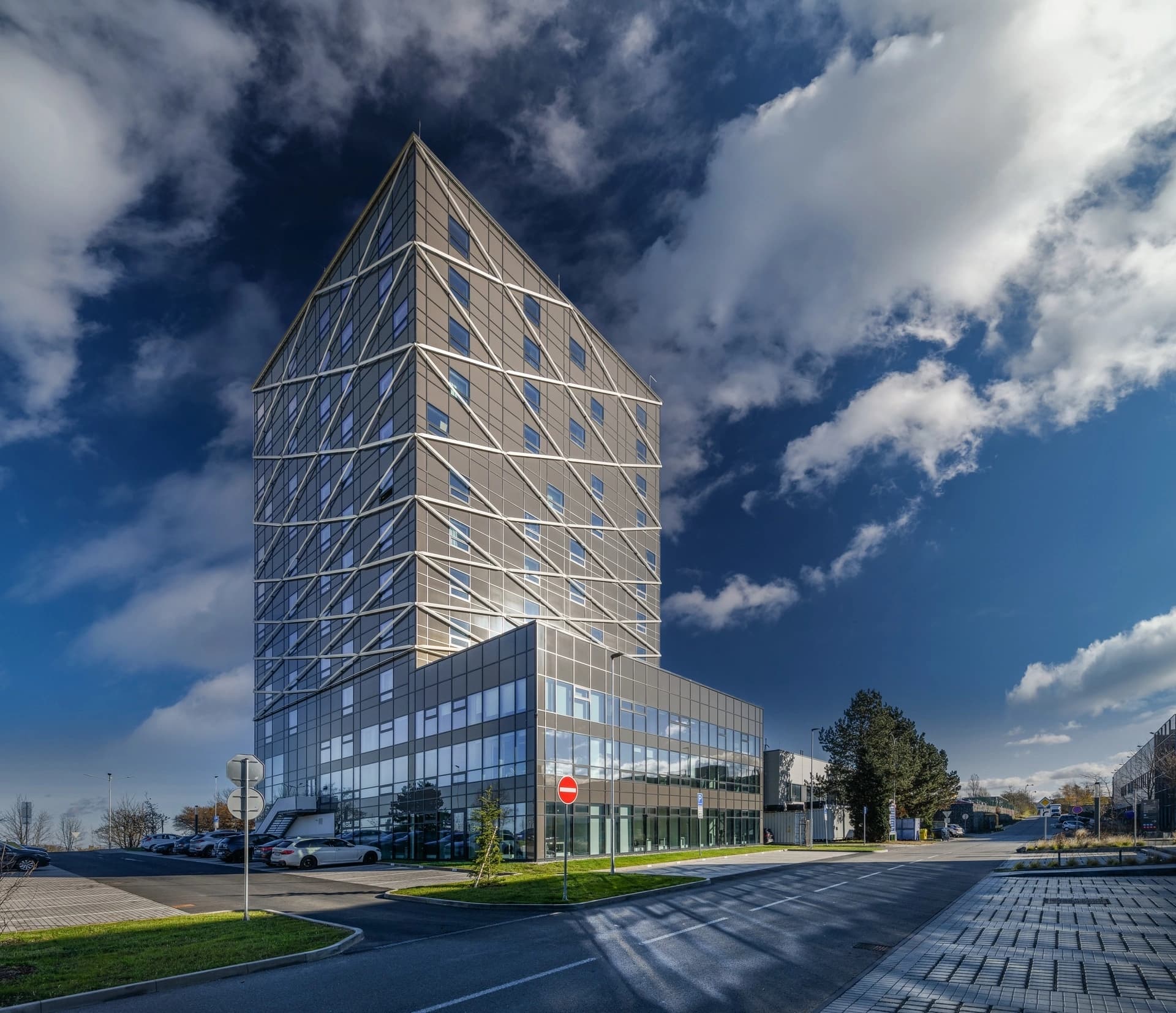 Bâtiment moderne et imposant avec une façade grise angulaire, un sommet triangulaire et un motif géométrique blanc, ponctué de fenêtres bleues réfléchissantes. Il est situé dans un environnement urbain avec des routes et un parking, sous un ciel bleu vif parsemé de grands nuages blancs.