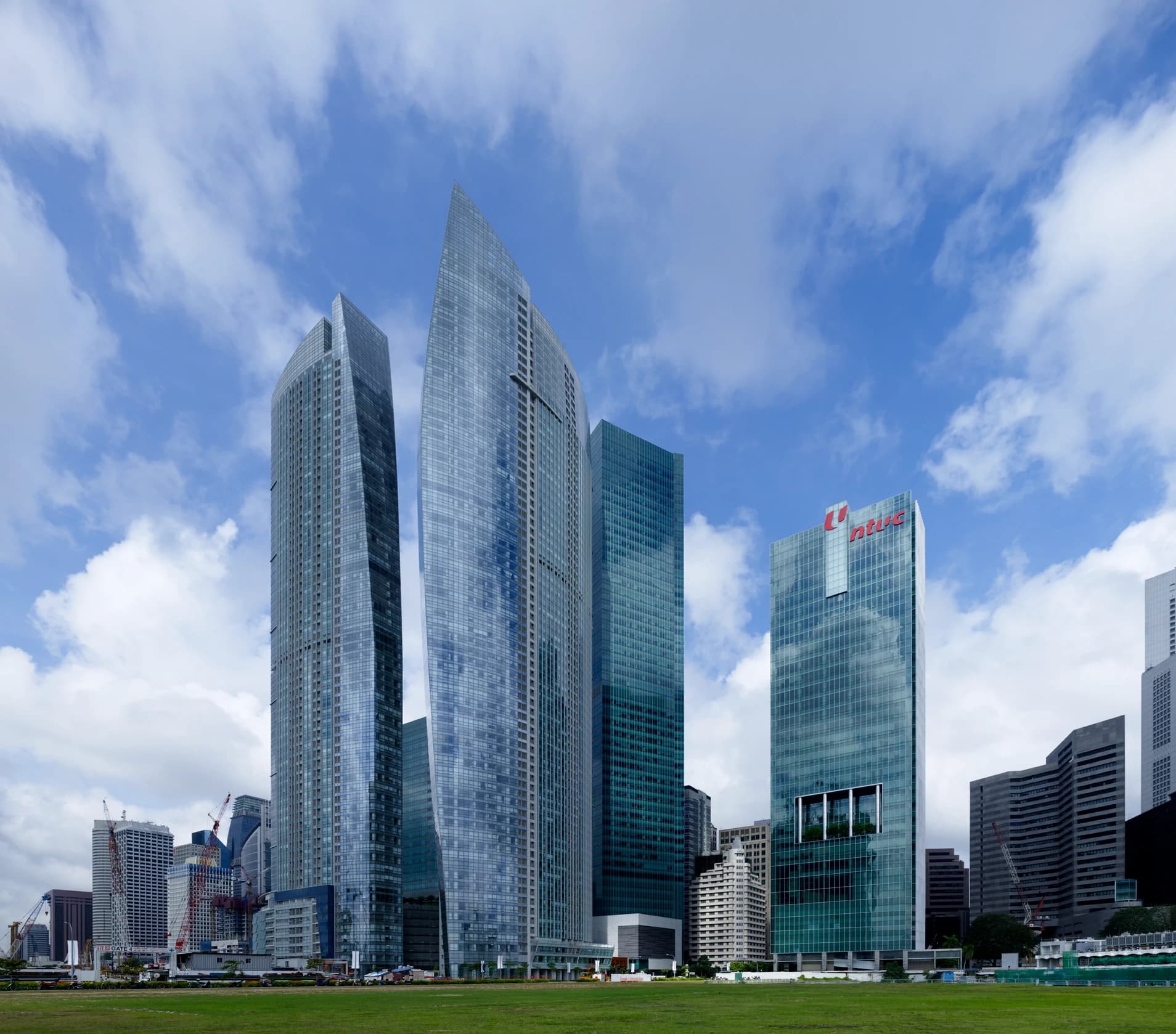 Des gratte-ciel modernes aux façades de verre bleuté s'élèvent au-dessus d'une pelouse verte, sous un ciel bleu lumineux parsemé de nuages blancs.