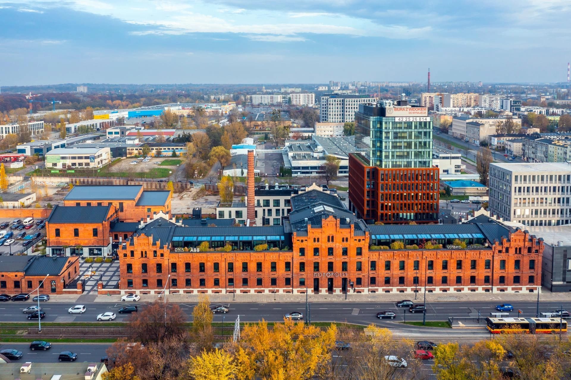 Vue aérienne d'un grand complexe urbain centré sur Monopolis, une ancienne usine en briques rouges avec toits sombres, entourée de bâtiments modernes, dont un gratte-ciel de verre et de métal, et d'arbres aux feuillages jaunes d'automne le long de routes animées avec un tram.