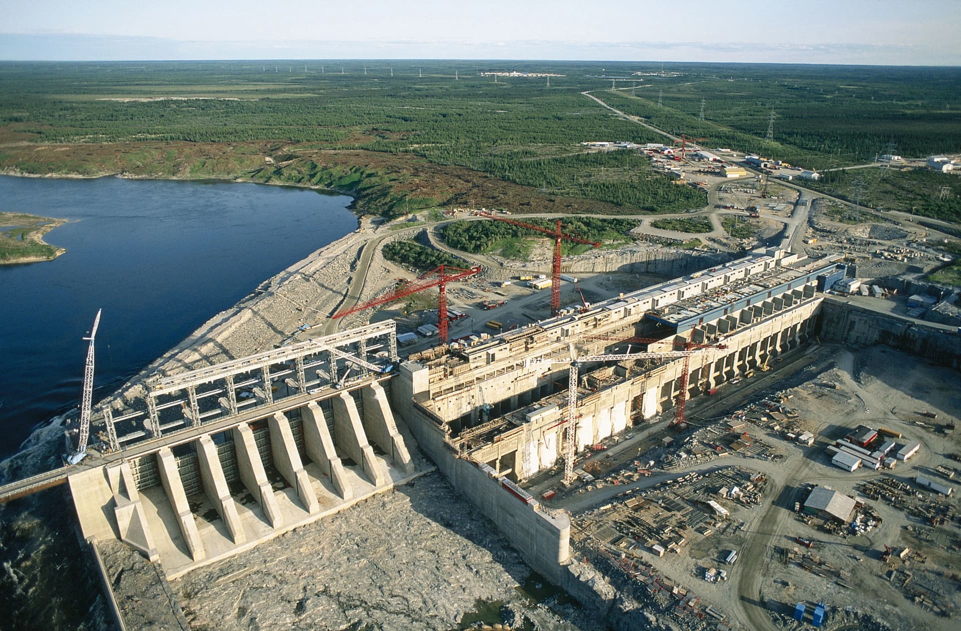 Vue aérienne d'un grand complexe de barrage hydroélectrique en béton gris, partiellement opérationnel avec de l'eau bleue s'écoulant, et en construction active avec de multiples grues rouges. Le vaste site industriel est entouré d'une dense forêt verte sous un ciel clair, avec des lignes électriques s'étendant à l'horizon.