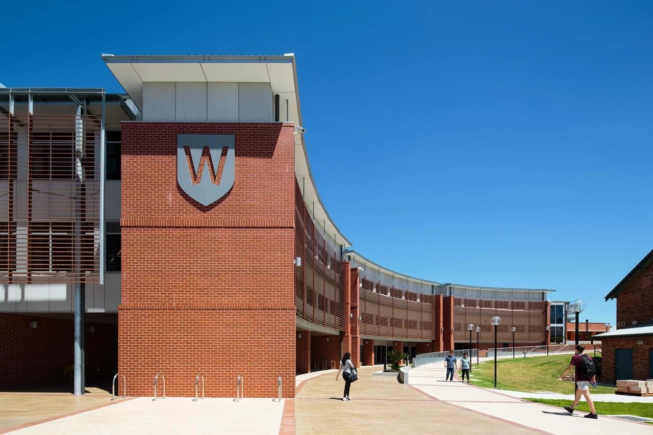 Un bâtiment moderne en briques rouges avec une façade incurvée et un logo 'W' sur un écusson, situé sur un campus ensoleillé où des étudiants marchent sur des allées.