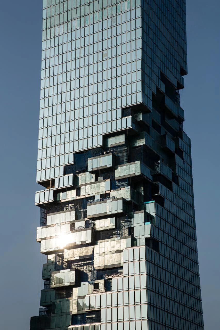 Un gratte-ciel moderne et imposant avec une façade en verre bleu-vert, caractérisée par des sections en saillie et en retrait qui donnent un effet pixelisé unique, sous un ciel bleu clair. Le soleil se reflète sur certaines fenêtres.