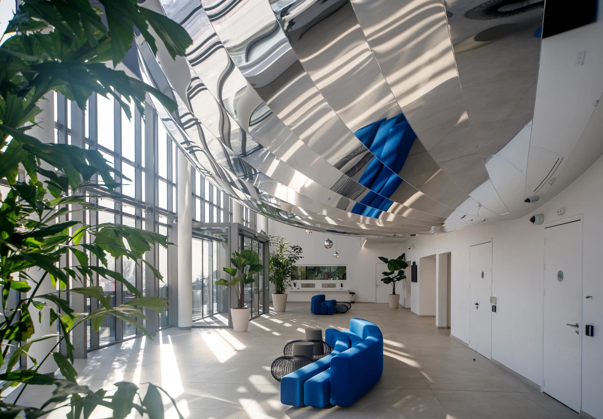 Un hall d'entrée moderne et lumineux présente un plafond ondulé et réfléchissant, de grandes fenêtres et des sièges modulaires bleus vifs. La lumière du soleil inonde le sol gris, éclairant des plantes en pot et créant des reflets dynamiques.