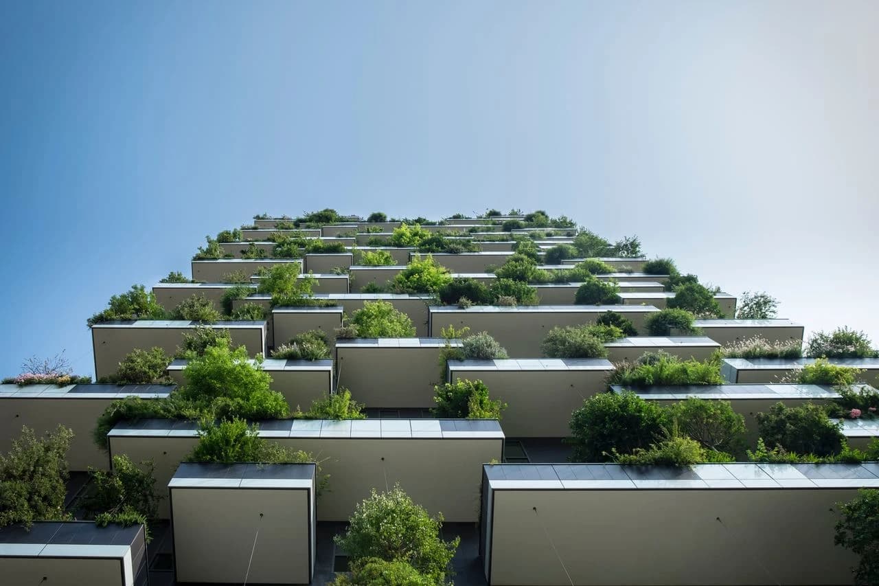 Vue en contre-plongée d'un bâtiment moderne où des balcons en cascade sont densément végétalisés d'arbres et d'arbustes verts. Cette architecture durable intègre la nature en milieu urbain, sous un ciel bleu clair.