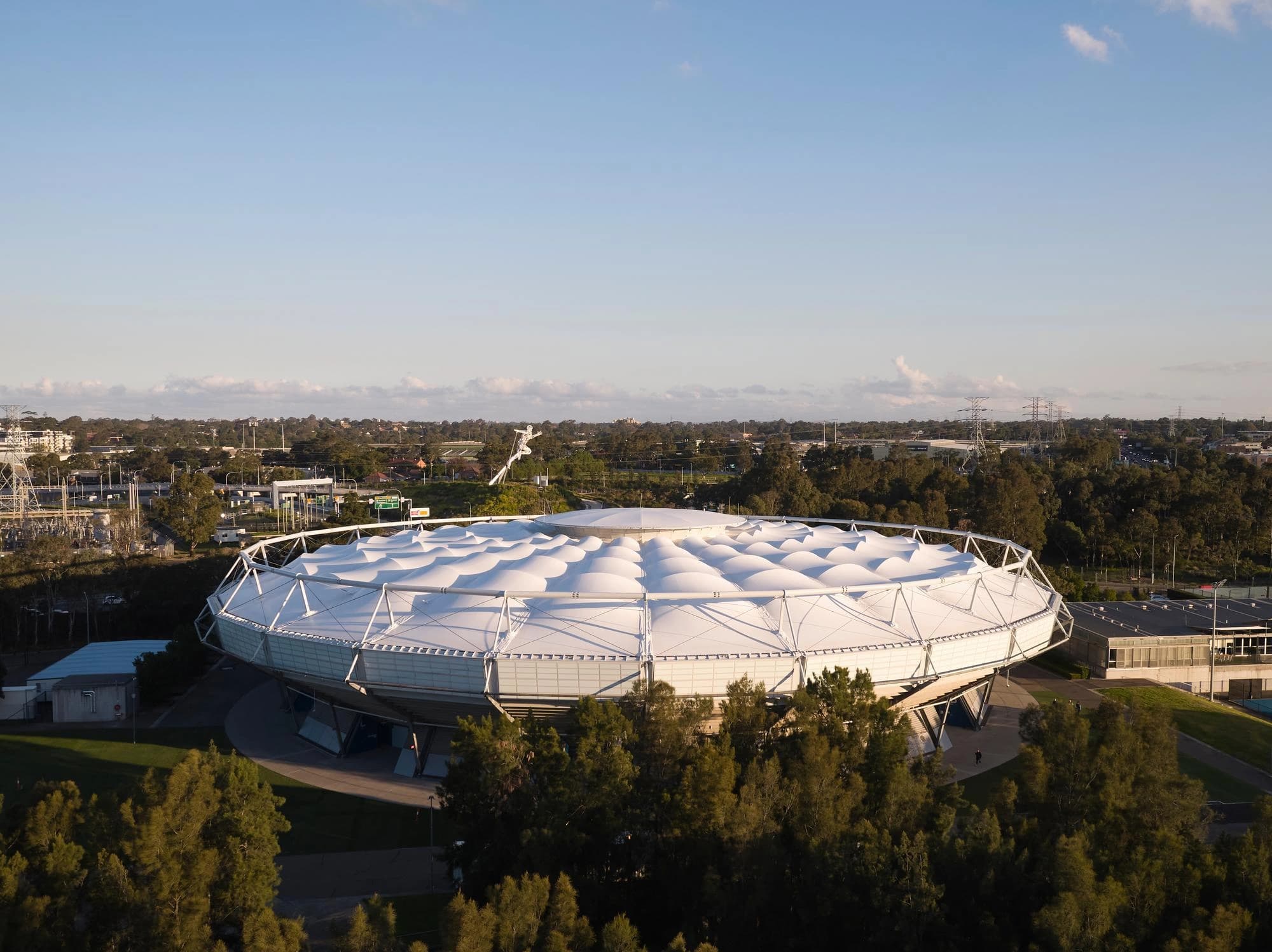 Vue aérienne d'un grand stade circulaire blanc à l'architecture moderne avec un toit en tissu gonflé multi-dômes, entouré d'arbres verdoyants et d'un paysage urbain sous un ciel bleu lumineux.