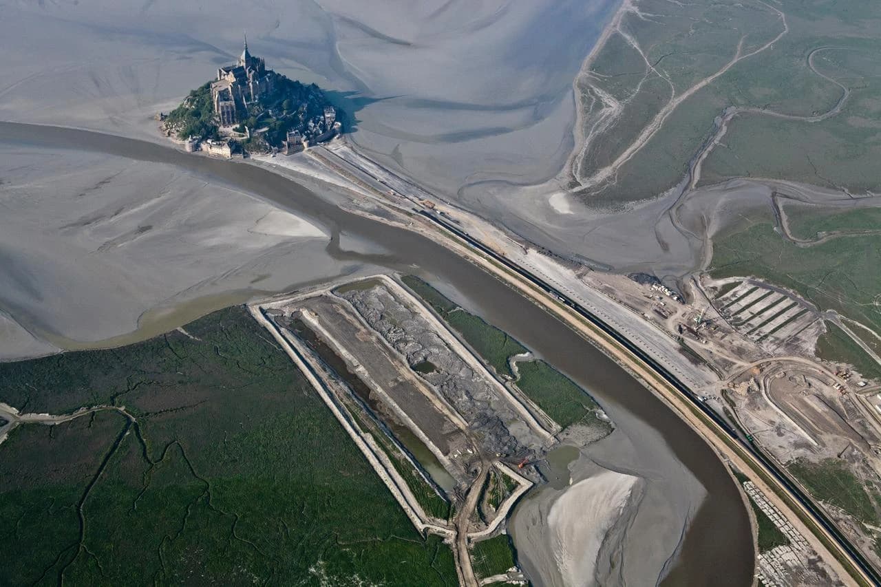 Vue aérienne du Mont Saint-Michel sur son îlot rocheux au milieu d'une baie de sable et de vasières grises, montrant d'importants travaux de dragage et la construction d'une nouvelle voie d'accès, bordée par des zones de verdure sombre.