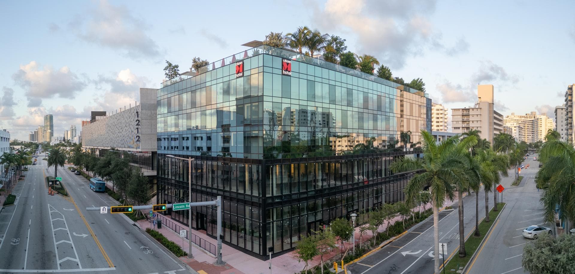 Un hôtel citizenM moderne à façade de verre, orné de logos rouges et de verdure sur le toit, se dresse à une intersection urbaine. Des palmiers bordent les rues animées, reflétant le ciel partiellement nuageux et les bâtiments environnants.