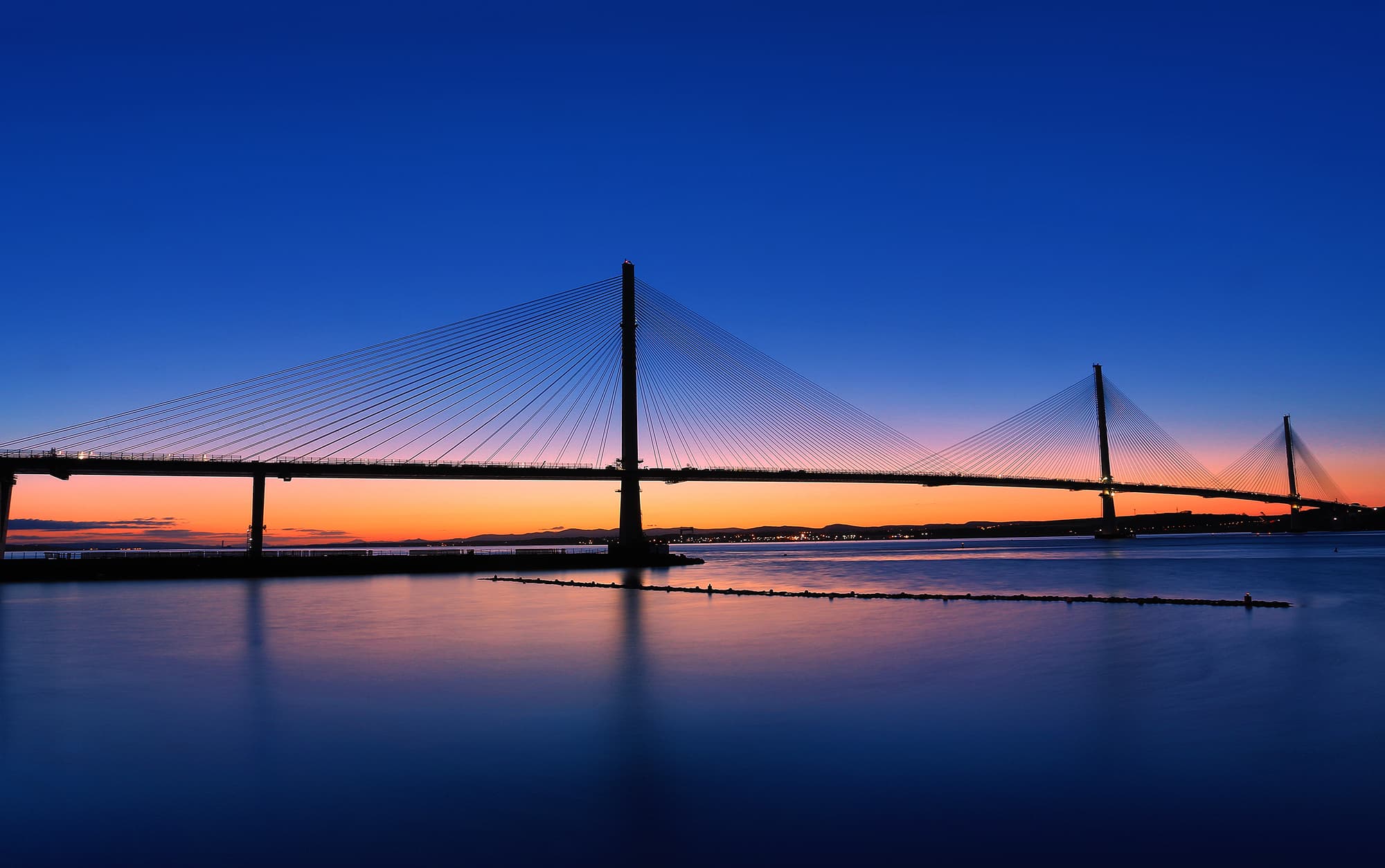 Pont à haubans en silhouette s'étendant sur l'eau calme au crépuscule, sous un ciel aux couleurs vives allant de l'orange à l'horizon au bleu nuit profond au zénith.