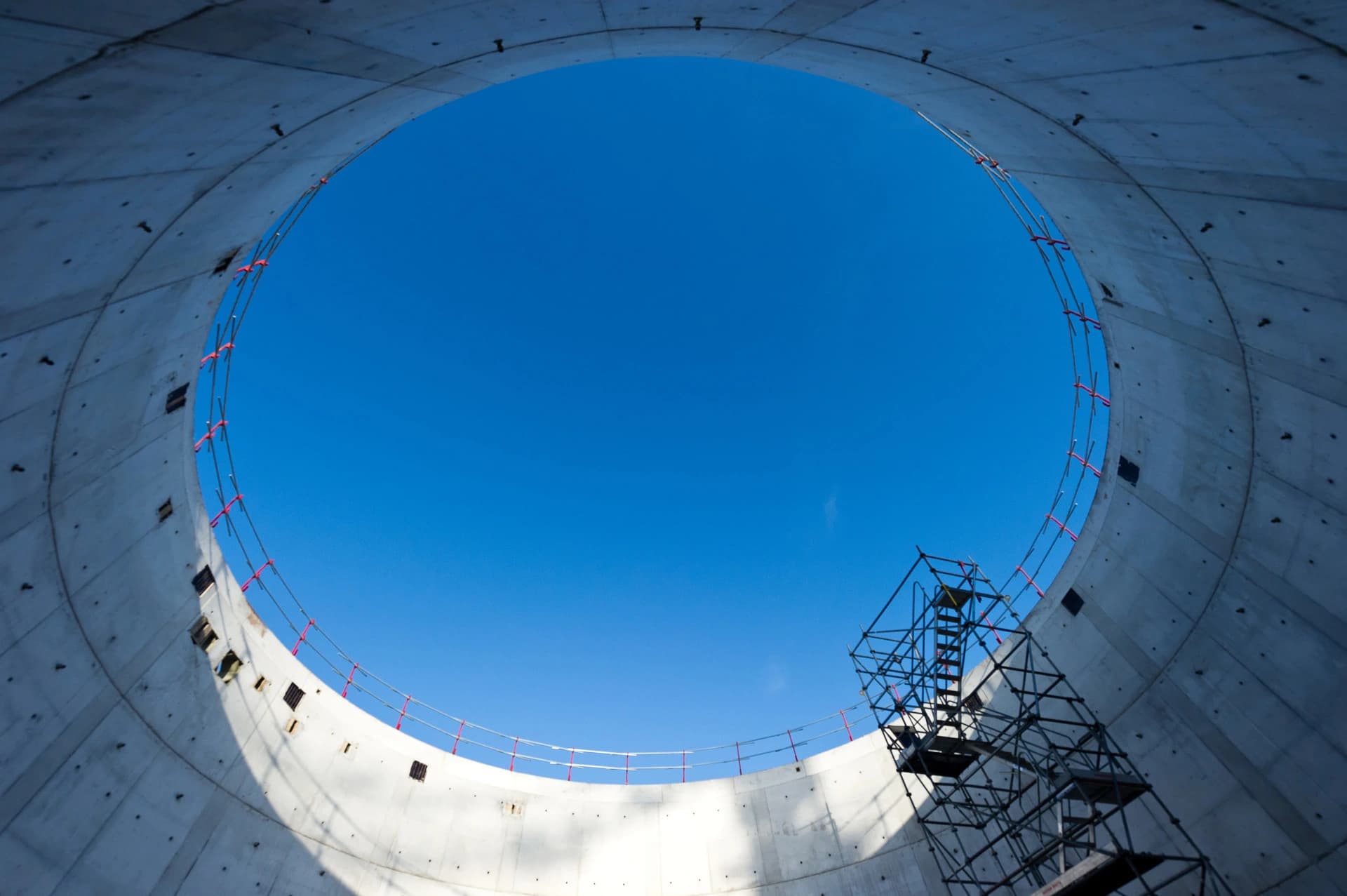 Vue de l'intérieur d'un cylindre en béton brut en construction, où un grand cercle de ciel bleu clair est visible au sommet, bordé par une balustrade rouge et un échafaudage métallique sombre.