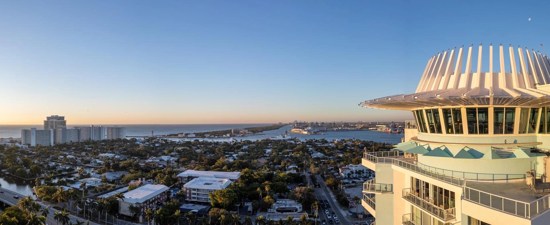 Vue panoramique d'une ville côtière au lever ou coucher du soleil, dominée par un bâtiment moderne blanc et bleu avec un toit circulaire distinctif au premier plan. Le paysage s'étend sur un port actif rempli de navires, des quartiers résidentiels verdoyants et l'océan à l'horizon sous un ciel dégradé.