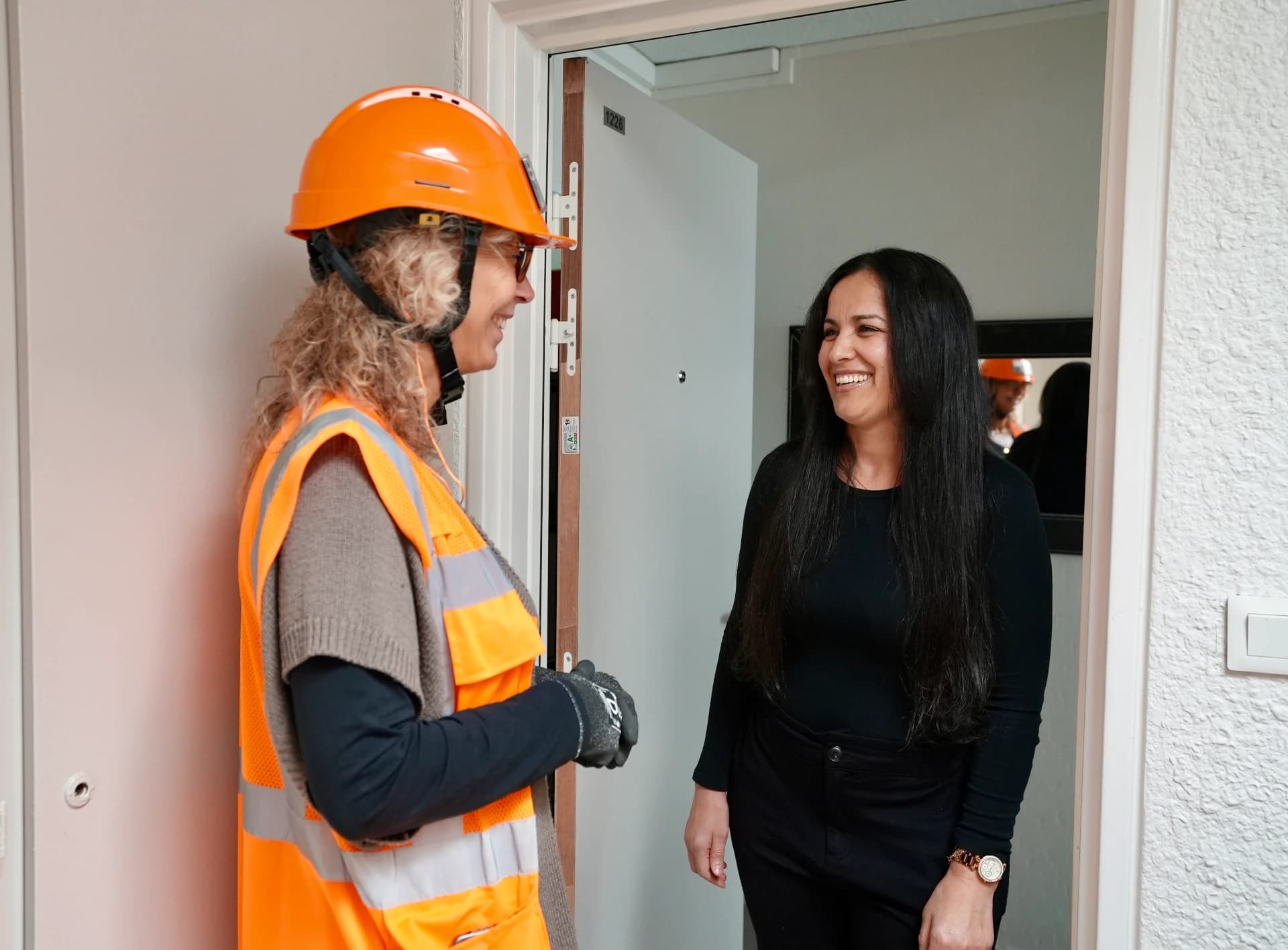 Une ouvrière souriante, portant un casque et un gilet orange fluo, échange avec une femme aux cheveux foncés qui sourit, devant la porte ouverte d'un appartement.