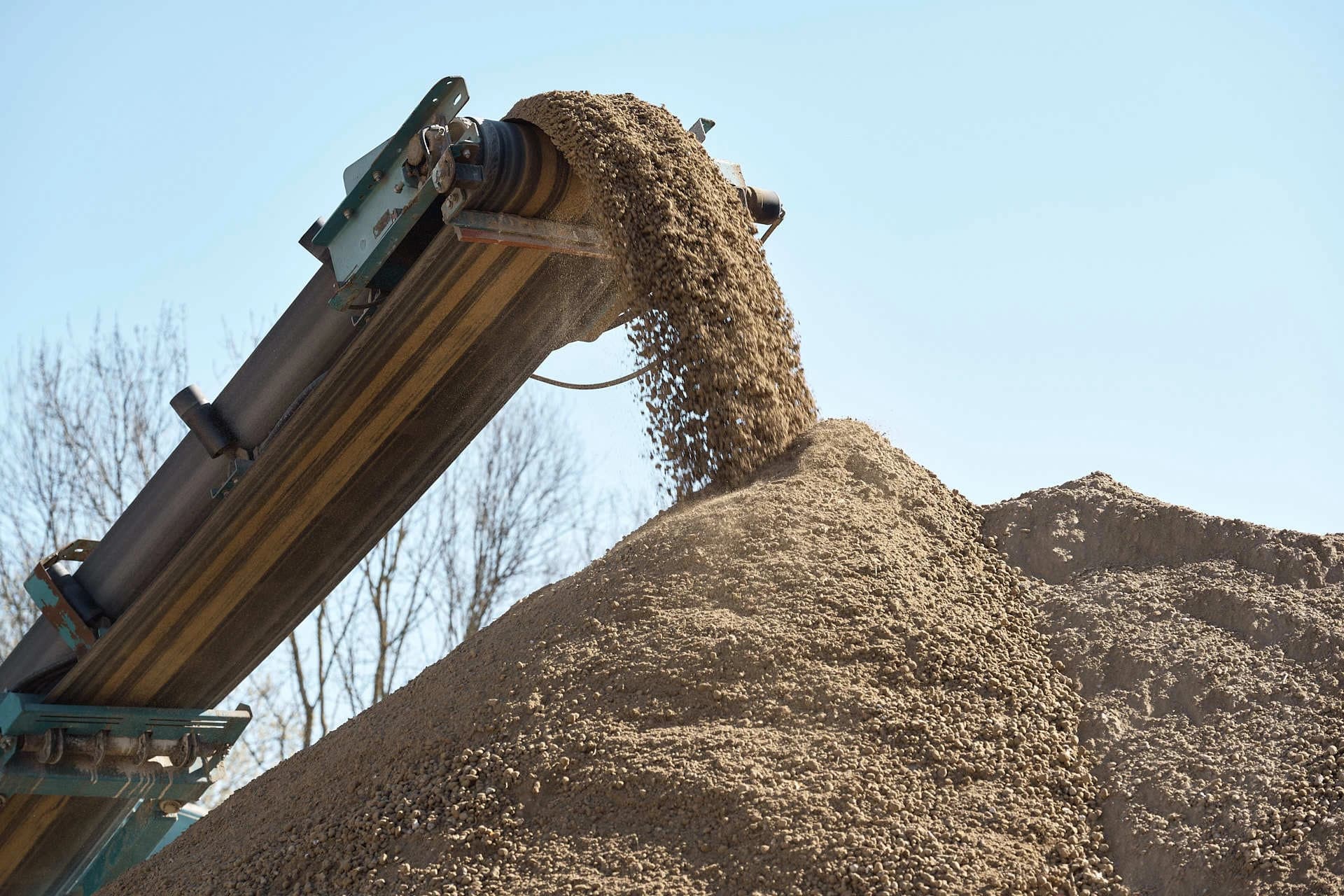 Un convoyeur industriel déverse activement du sable et du gravier clairs sur une grande pile, sous un ciel bleu dégagé.