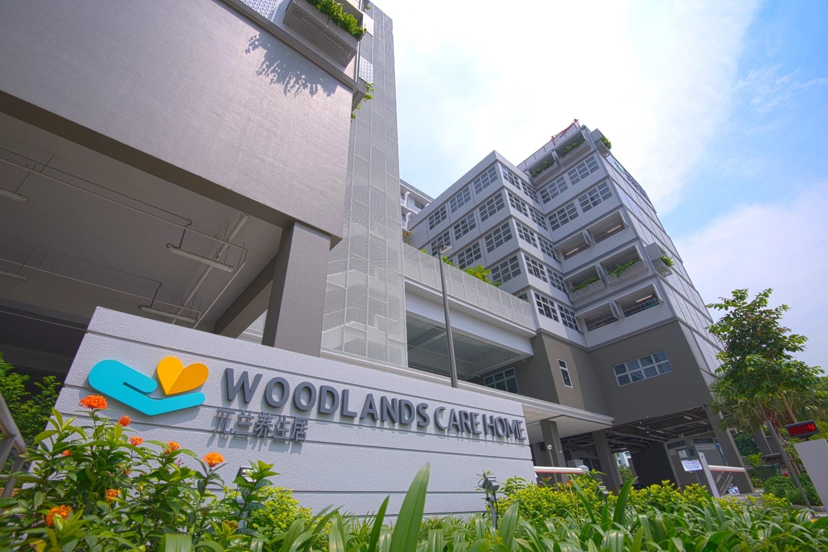 A modern, multi-story grey building, Woodlands Care Home, features its prominent sign with a teal hand and yellow heart logo, surrounded by vibrant green foliage and orange flowers under a bright blue sky.